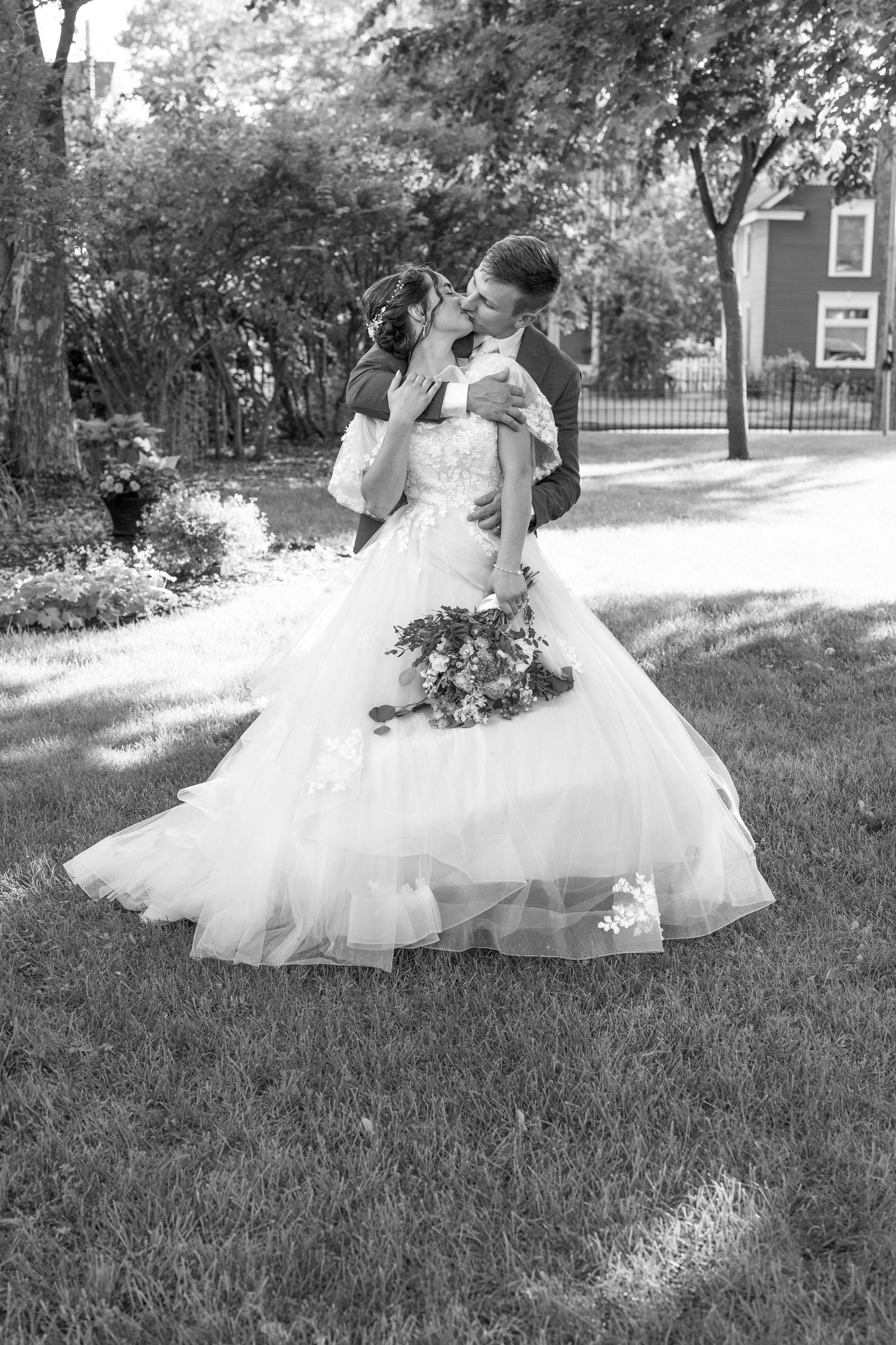 Black and white photo of a bride and groom sharing a kiss in a lush garden setting.