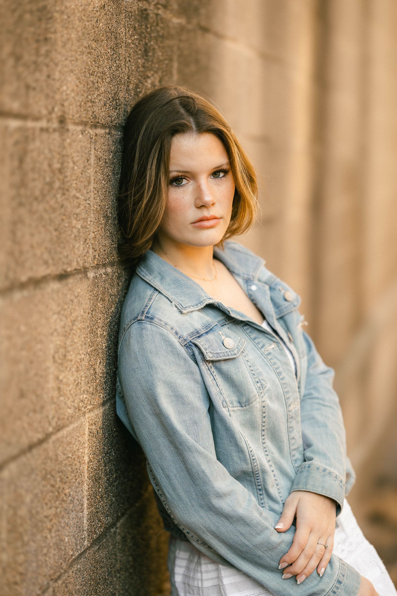 Young woman in denim jacket leaning against stone wall, casual portrait photography.