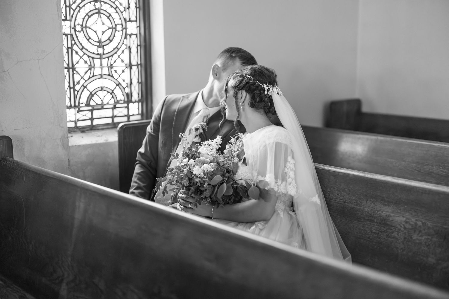 Black and white photo of bride and groom sharing a kiss in a church pew with stained glass.