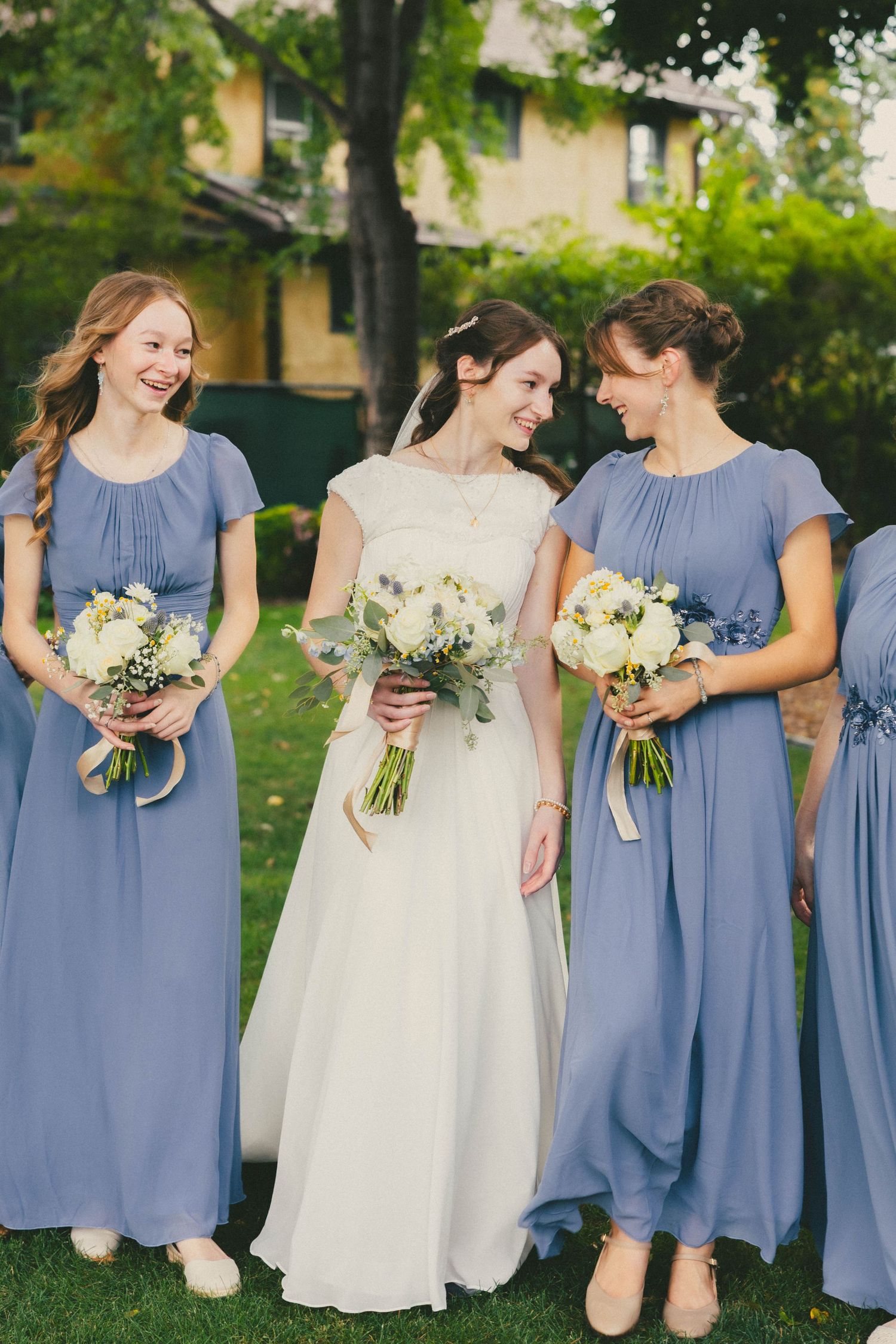 Bride in white gown stands with two bridesmaids in periwinkle blue dresses holding white floral bouquets outdoors.