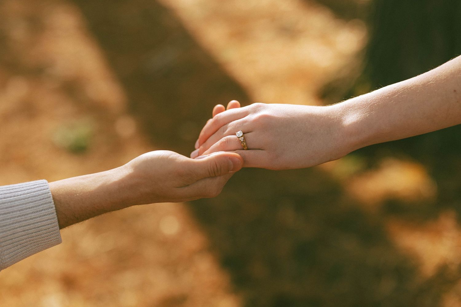 Two people gently holding hands outdoors, one wearing a diamond engagement ring, with warm autumn bokeh background.