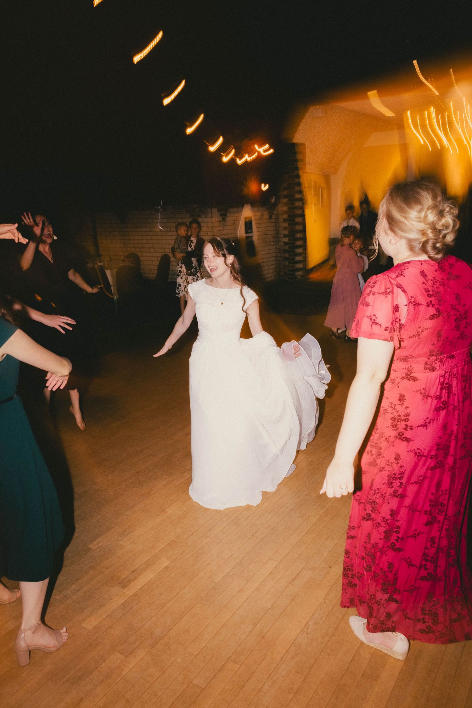 Bride in white gown dancing joyfully with guests on wooden dance floor at elegant wedding reception.