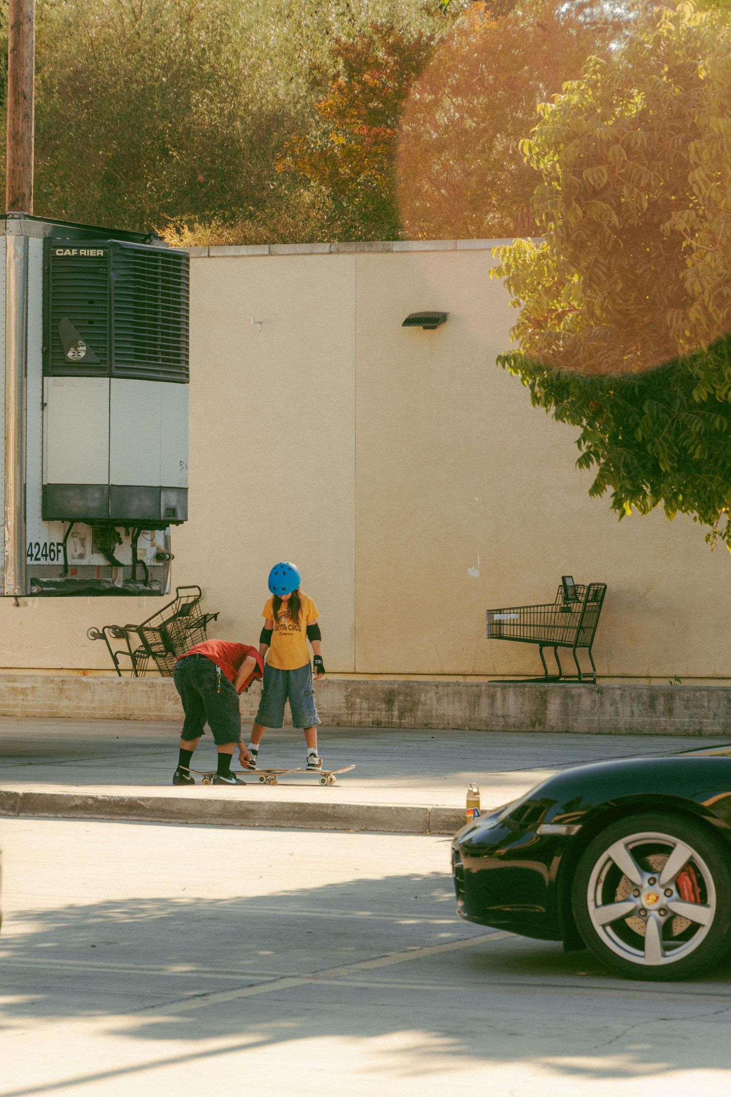 Two children skateboarding in sunny parking lot near shopping carts with Porsche car and autumn trees.