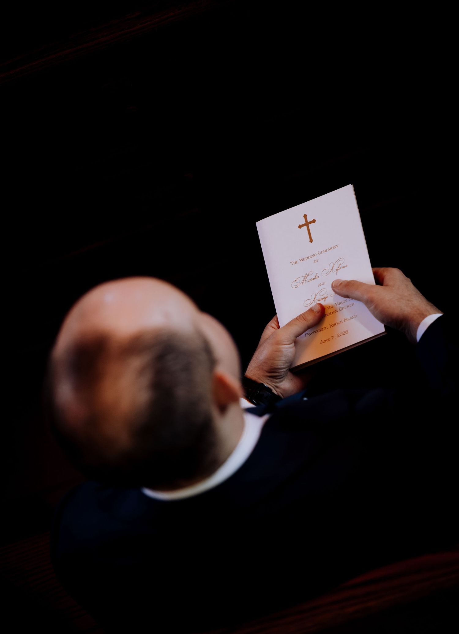 Man in suit reading a religious church program booklet with a gold cross during a ceremony.