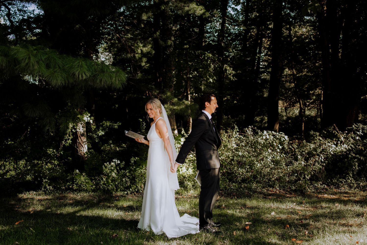 A wedding couple stands back-to-back in a sunlit garden setting.
