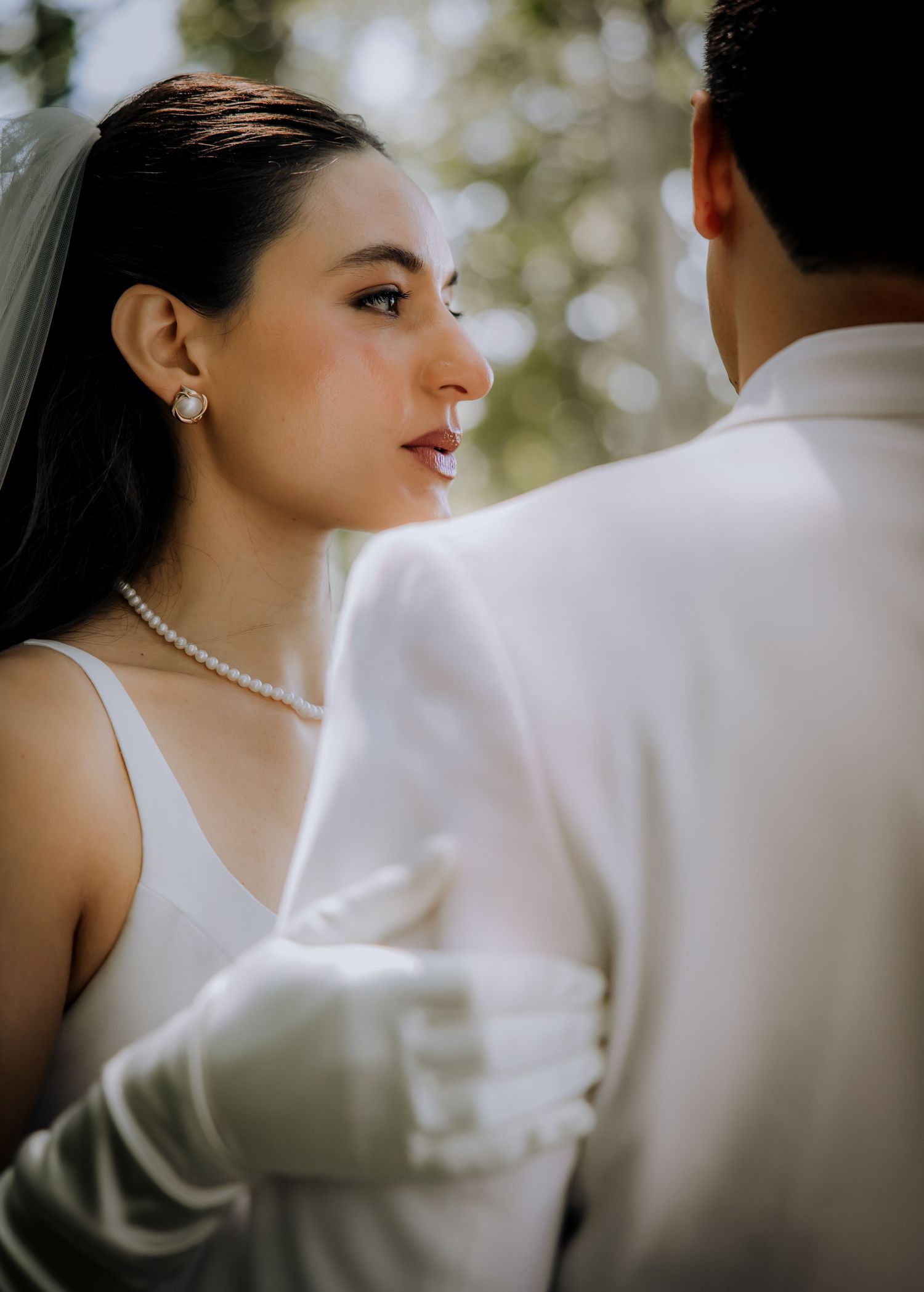 Bride in white dress and pearl jewelry gazes at groom in white suit during outdoor wedding ceremony.Editorial and Cinematic Wedding photos