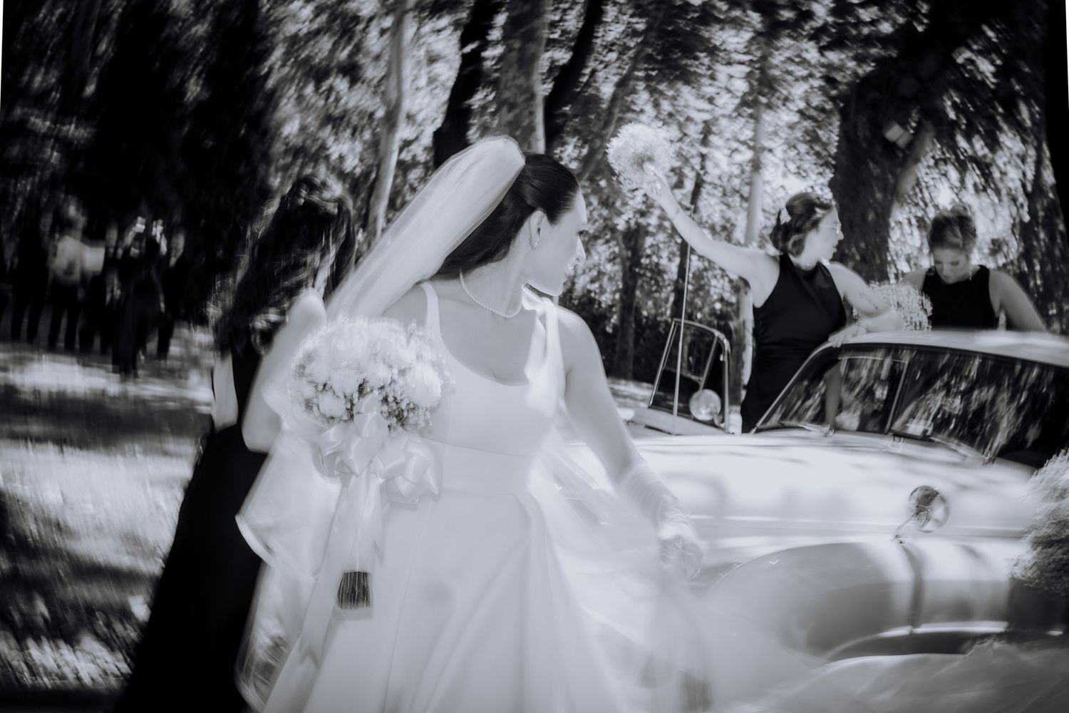 A black and white photo of a bride in a flowing dress with bouquet near a vintage car.