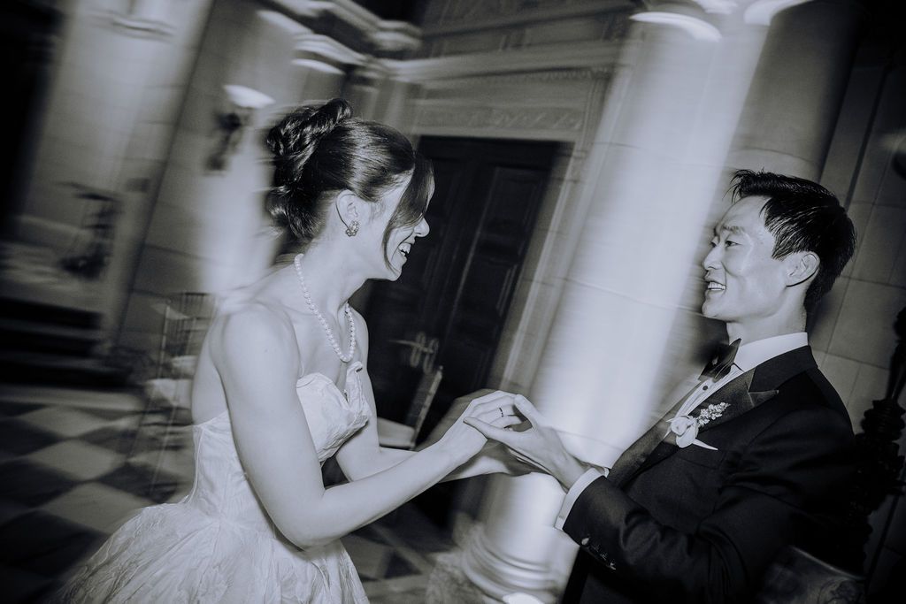 Black and white photo of a smiling couple exchanging rings at their wedding ceremony near elegant columns.