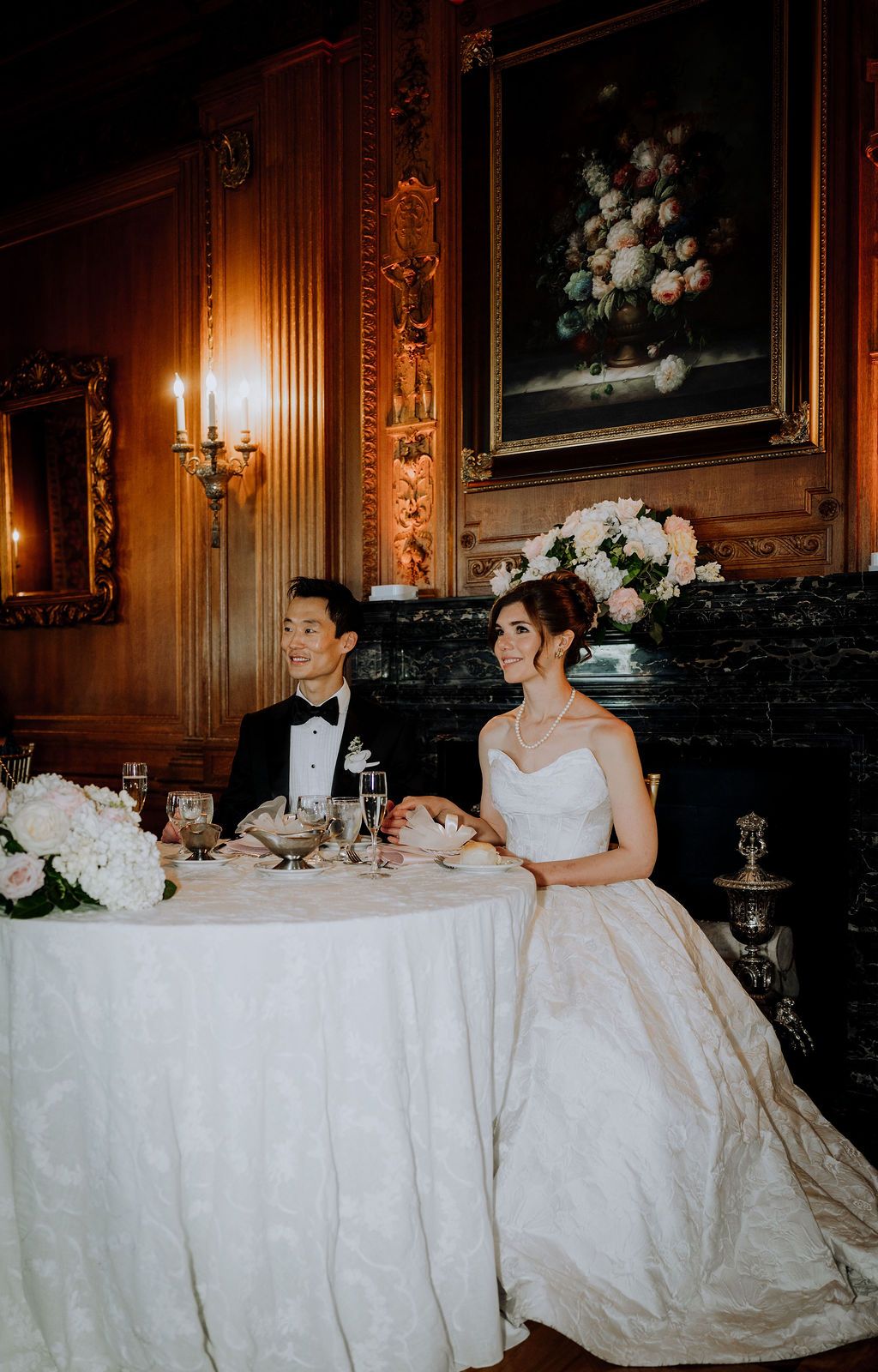 Bride and groom sit at sweetheart table in elegant wood-paneled reception hall with floral painting and candlelit sconces.