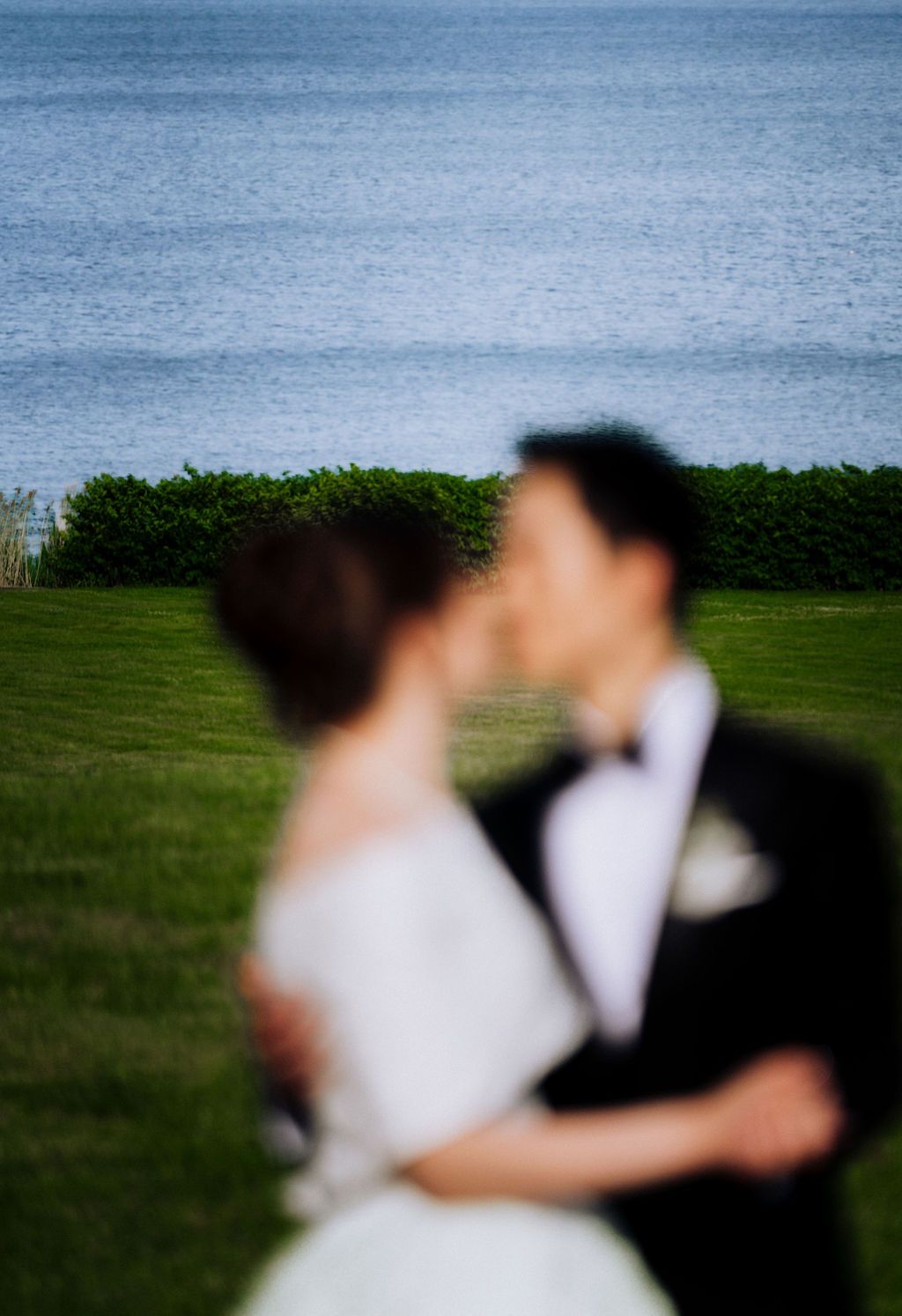 Blurred bride and groom sharing a kiss on green lawn with ocean and hedge backdrop on wedding day.