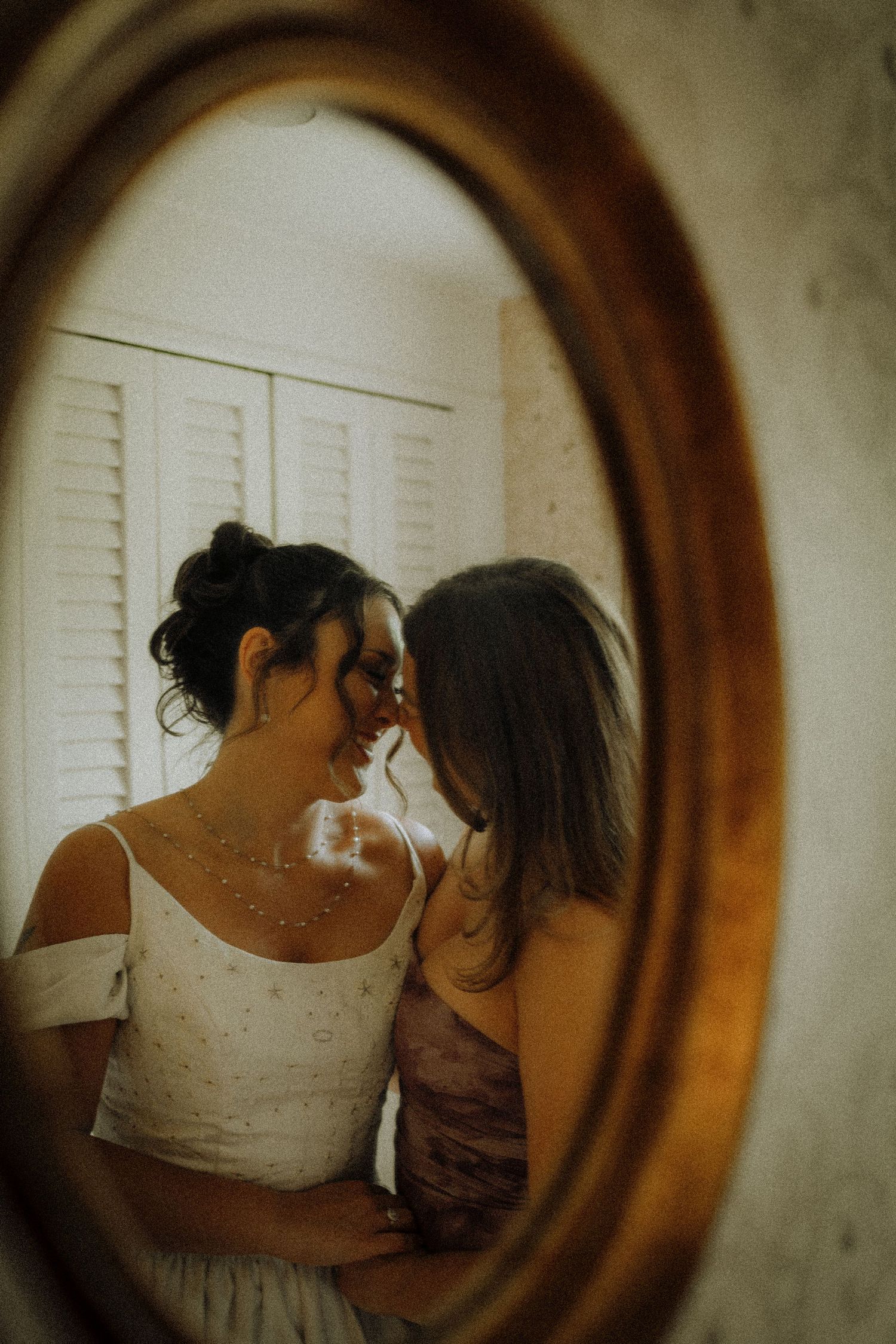 Two women share an intimate moment reflected in a vintage oval gold mirror, one wearing a white bridal gown.