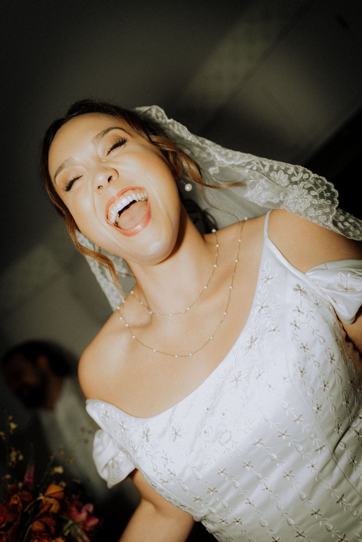 Joyful bride laughing in white embroidered gown with veil and pearl necklace on her wedding day.