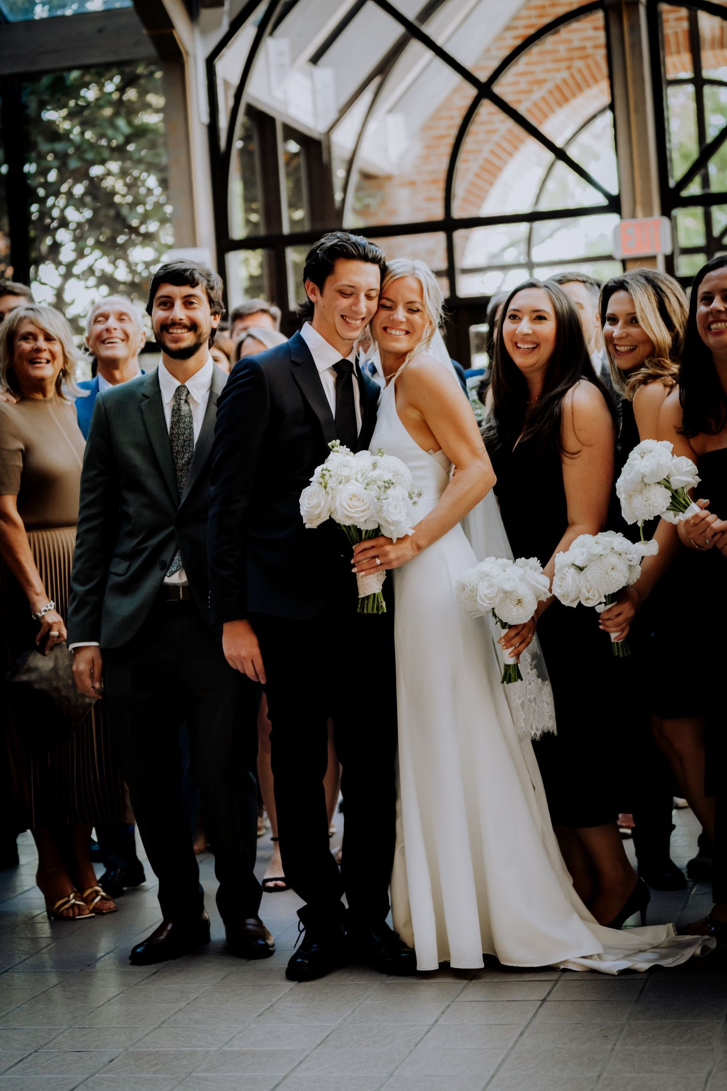 Wedding party poses together with white bouquets in an elegant venue with arched windows.