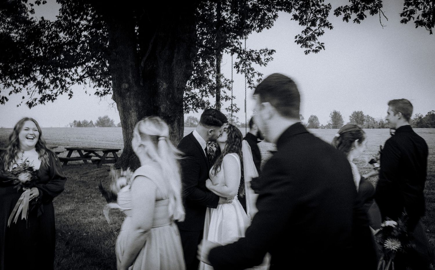 Black and white photo of wedding guests mingling under trees during outdoor reception.