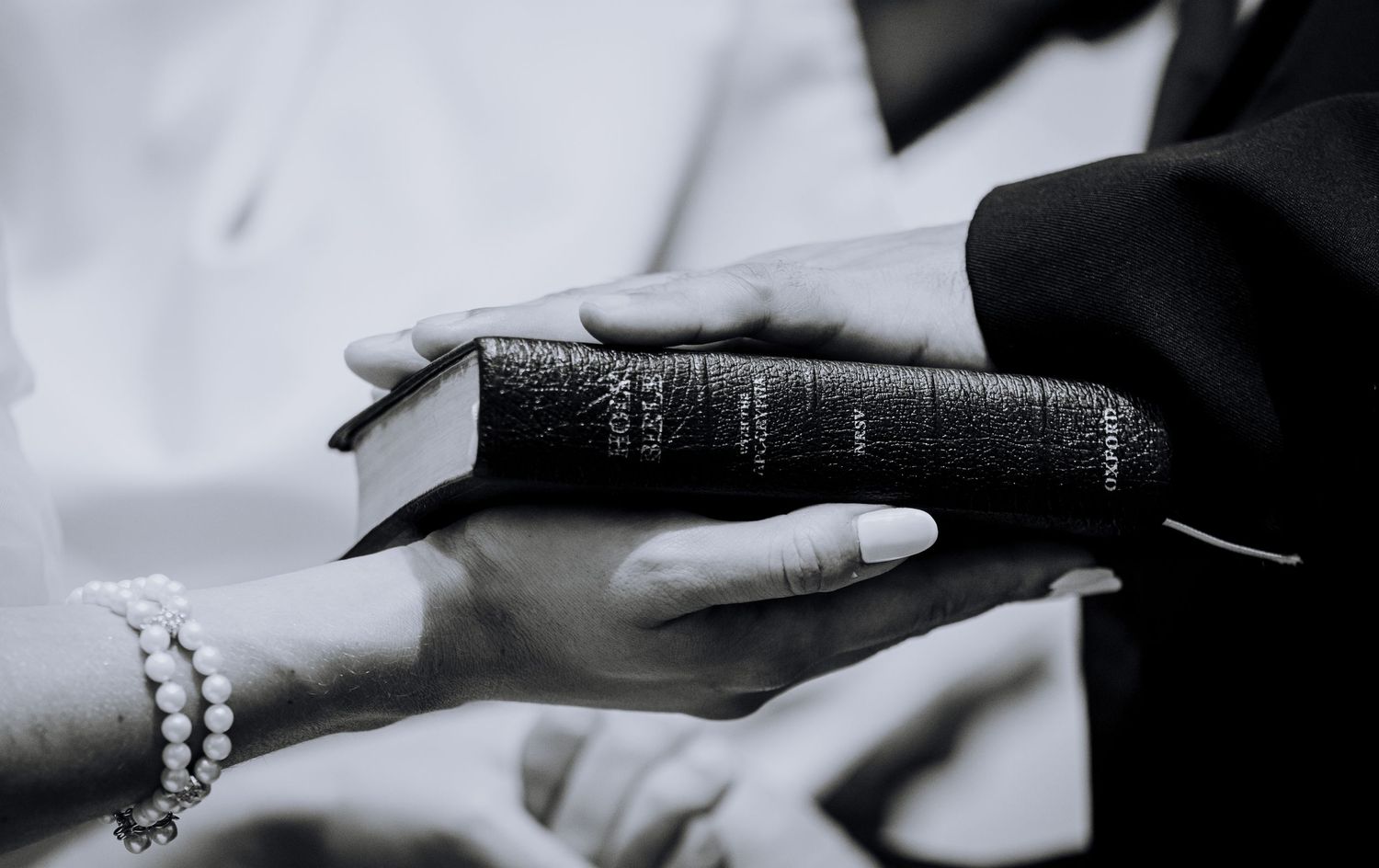 Close-up black and white photo of hands holding a Holy Bible, with a pearl bracelet visible.