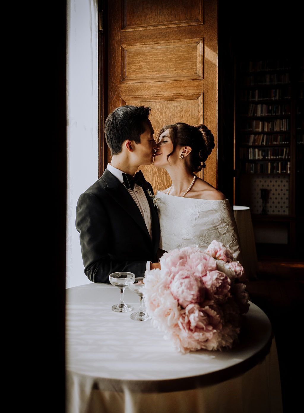 Newlyweds share a tender kiss beside a table with pink peony bouquet and champagne glasses.