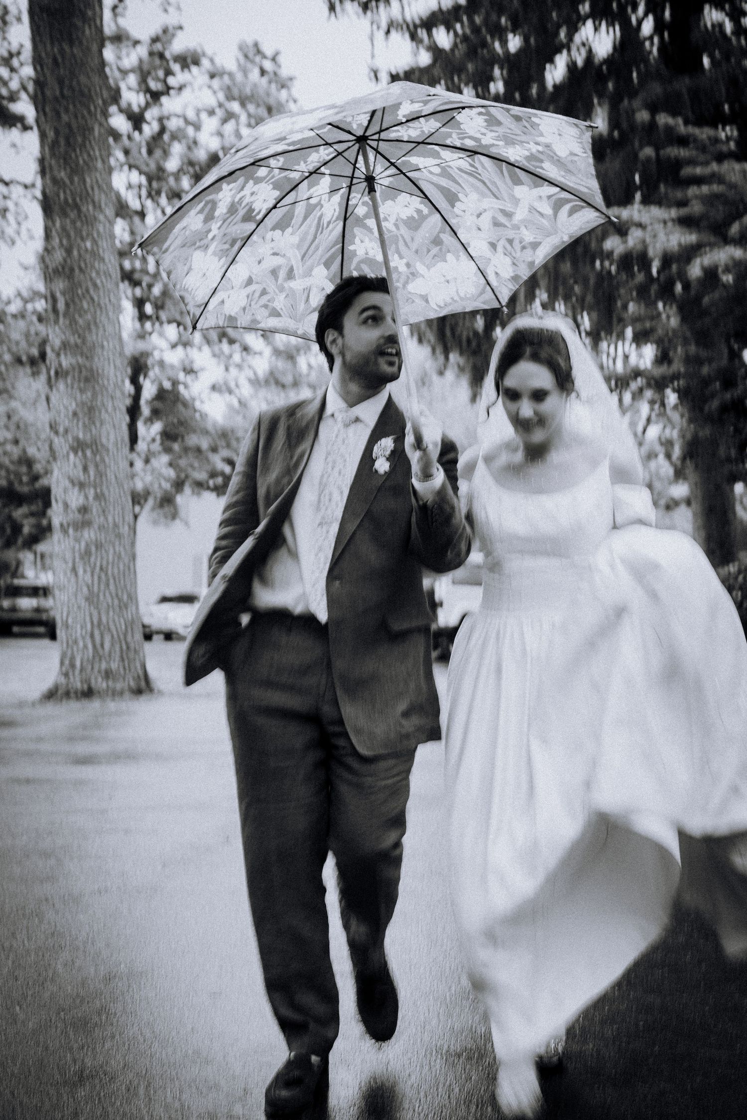 Black and white photo of a bride and groom walking under a clear umbrella on their rainy wedding day.