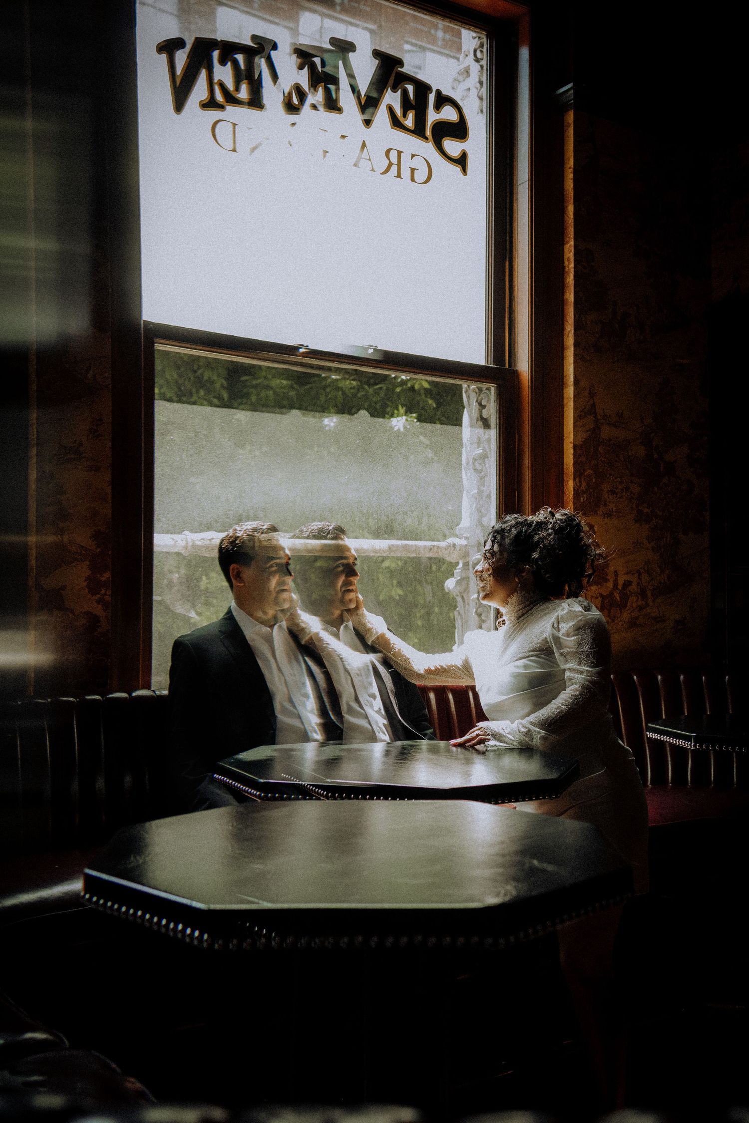 Bride and groom sit intimately at a dark wooden table by a window at Seven Grand bar, sharing a tender moment.
