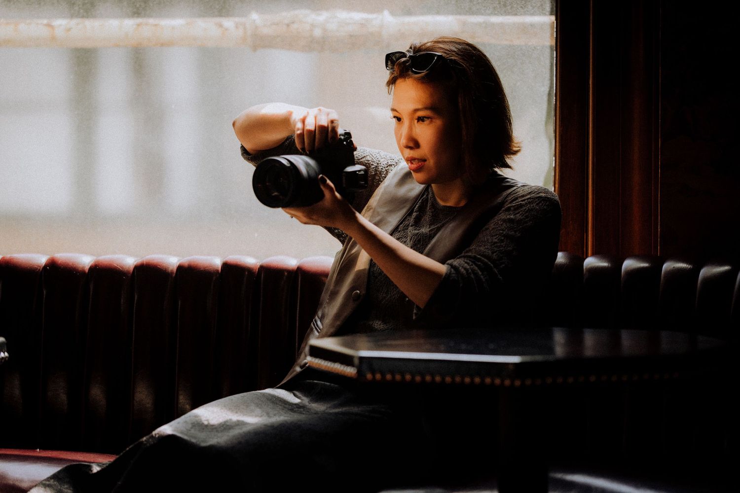 Woman photographer examining camera while seated by window in dimly lit room with leather seating.