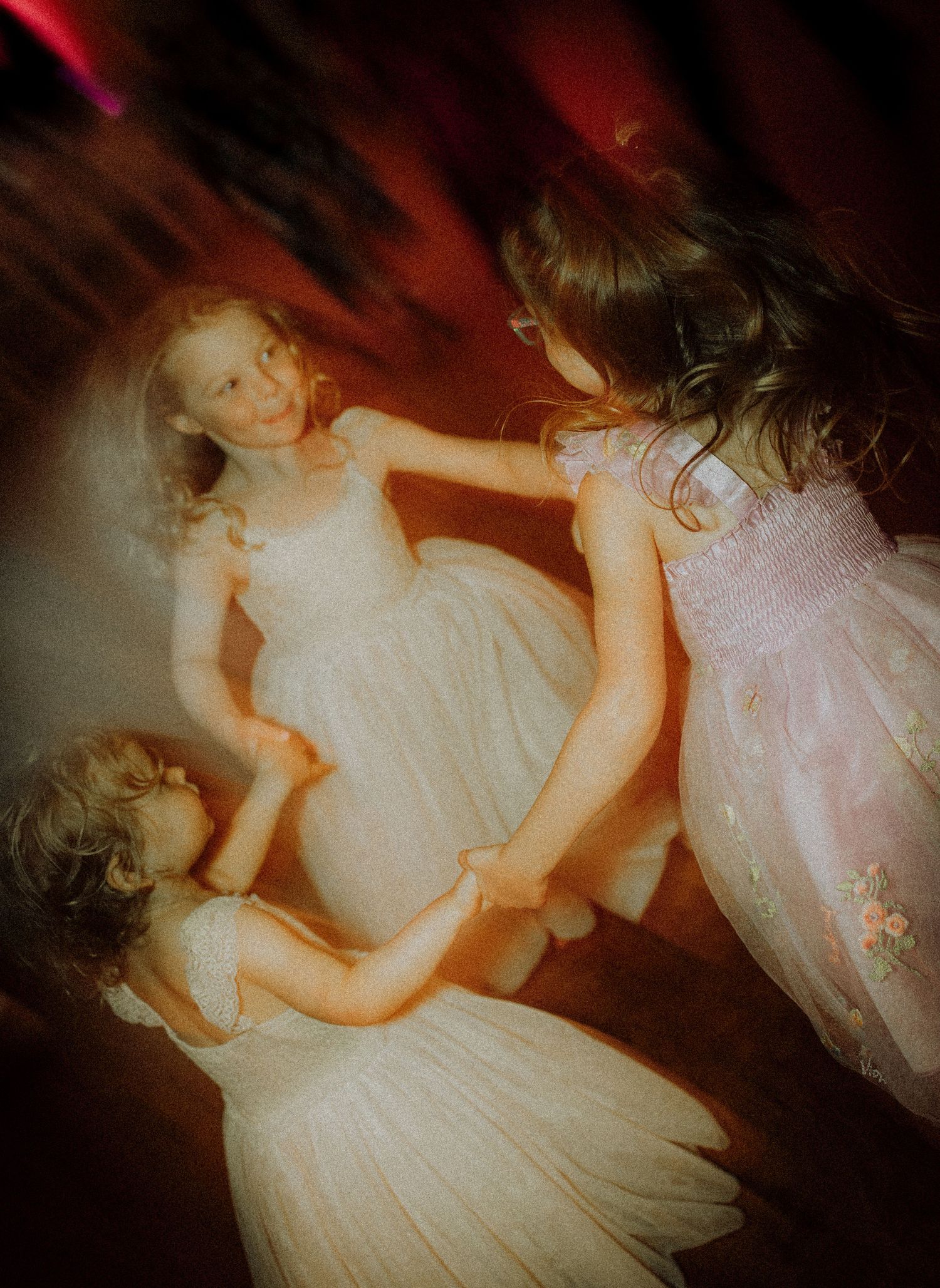 Three young girls in white dresses holding hands and dancing together in a circle at a formal event.