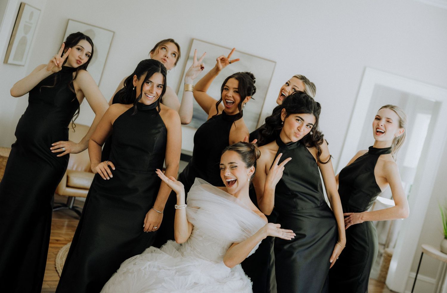 Bride in white gown poses with bridesmaids in black halter dresses, laughing and making fun gestures indoors.