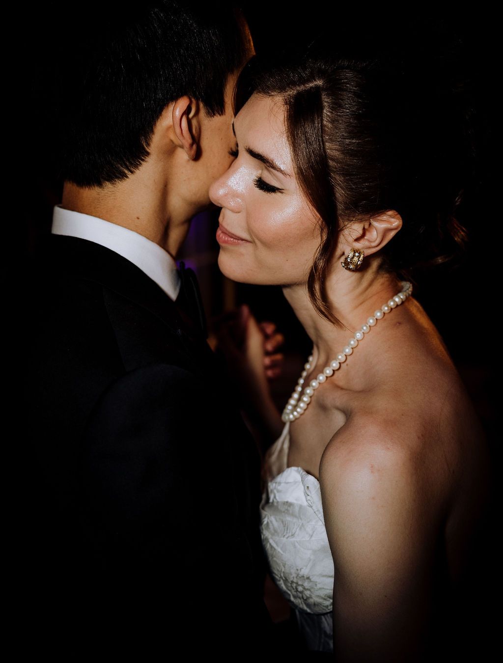 Bride in pearl necklace and strapless gown shares an intimate moment with groom in black suit at wedding reception.