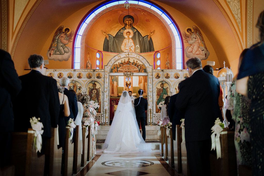 Wedding ceremony in ornate Orthodox church with bride in white gown walking down aisle toward golden iconostasis.