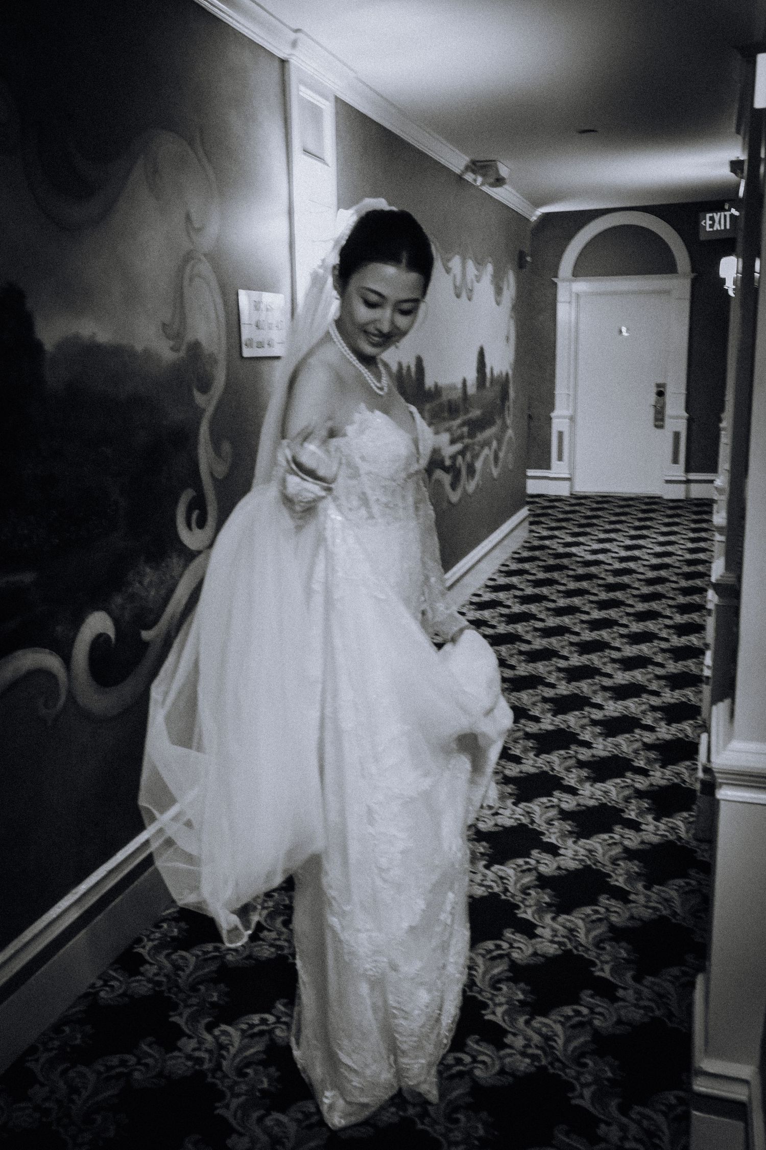 Black and white photo of a smiling bride in a lace wedding gown and veil standing in an elegant hotel hallway.