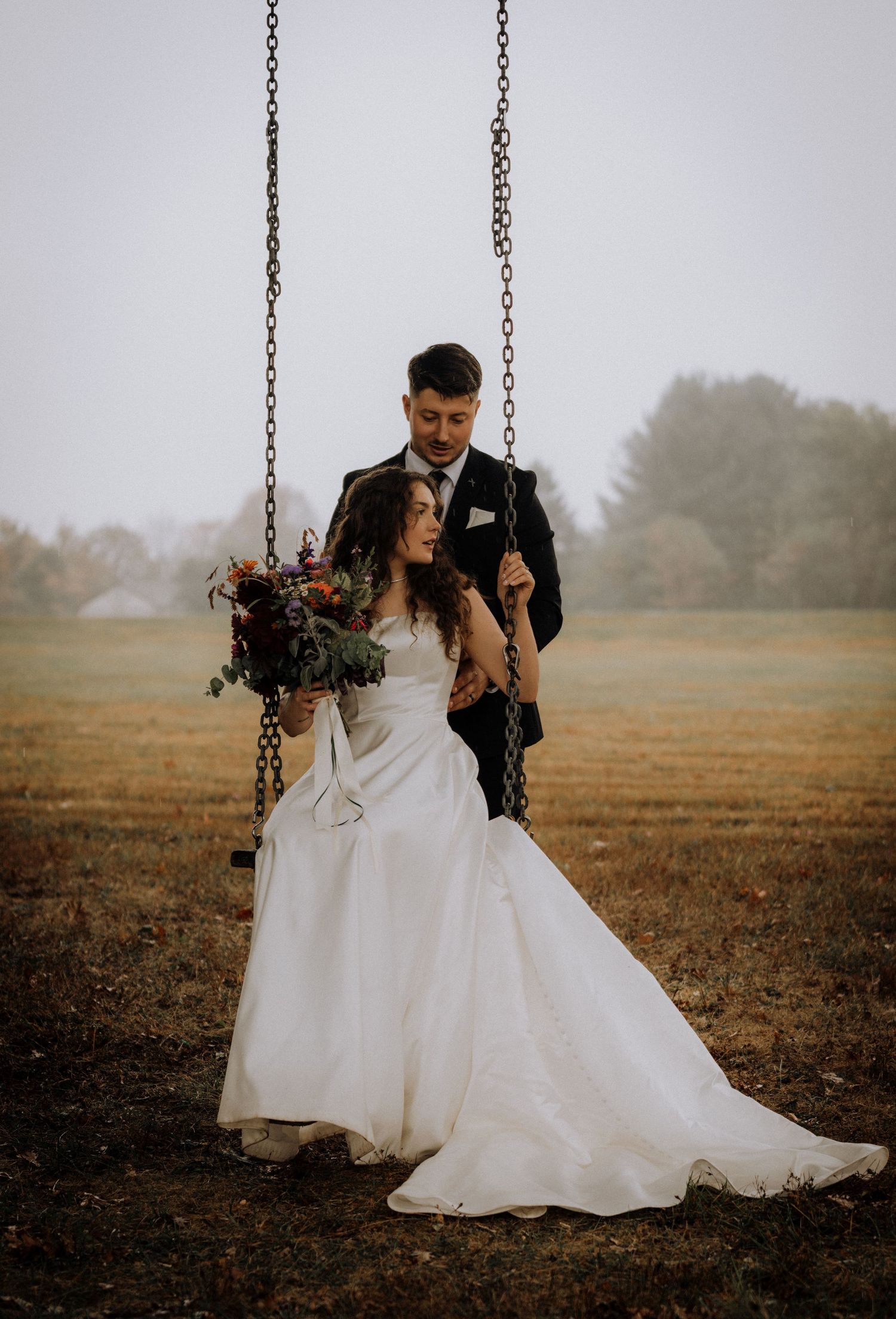 Romantic wedding portrait of couple on swing in misty autumn field.