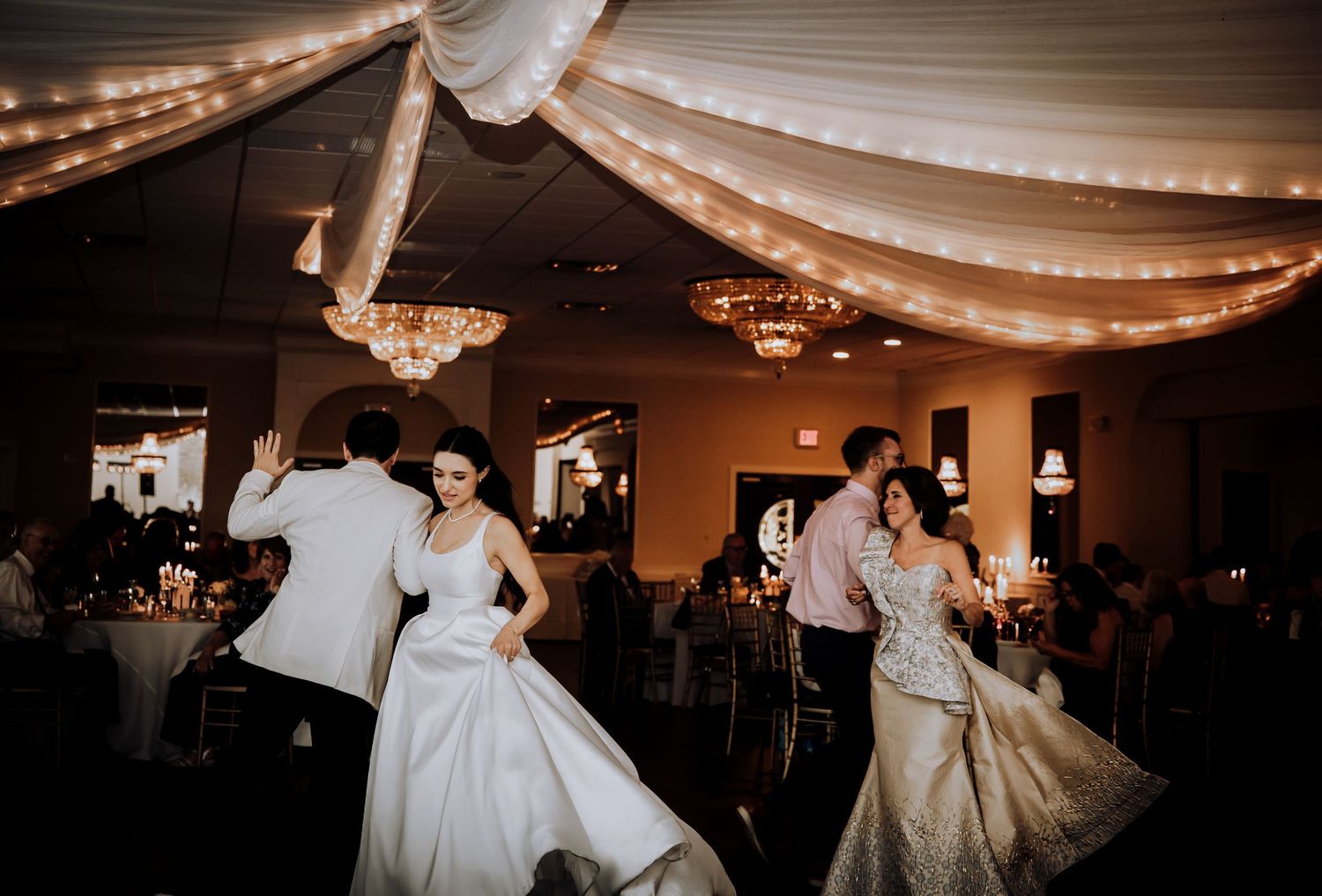 Couples dancing in elegant white dresses under draped fabric and string lights at wedding reception.