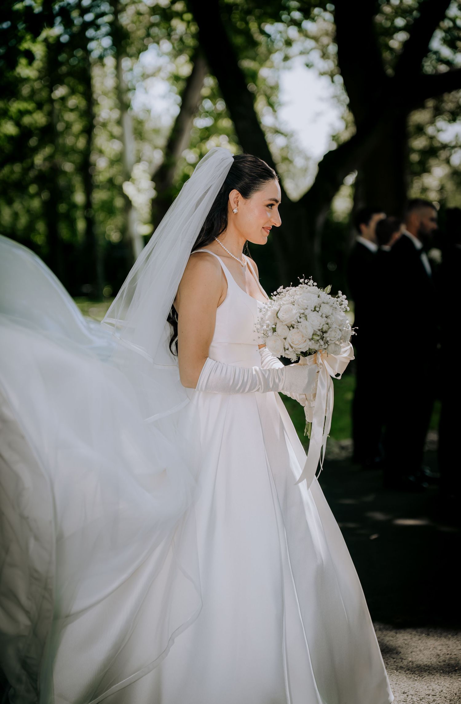 A bride in a sleeveless white gown and long gloves holds a white bouquet in an outdoor garden setting.