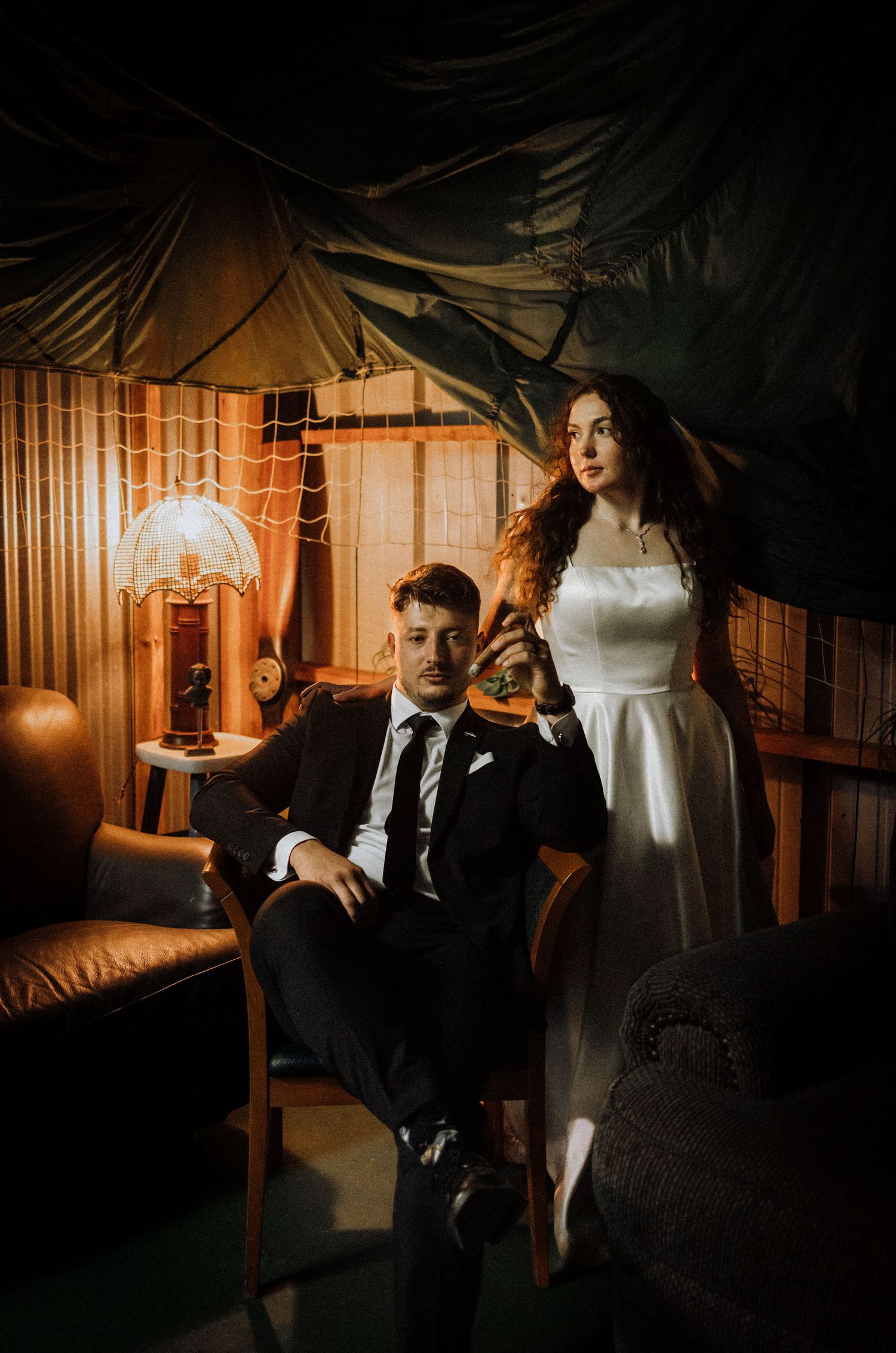 Wedding dress hangs in background while couple poses in dimly lit room.