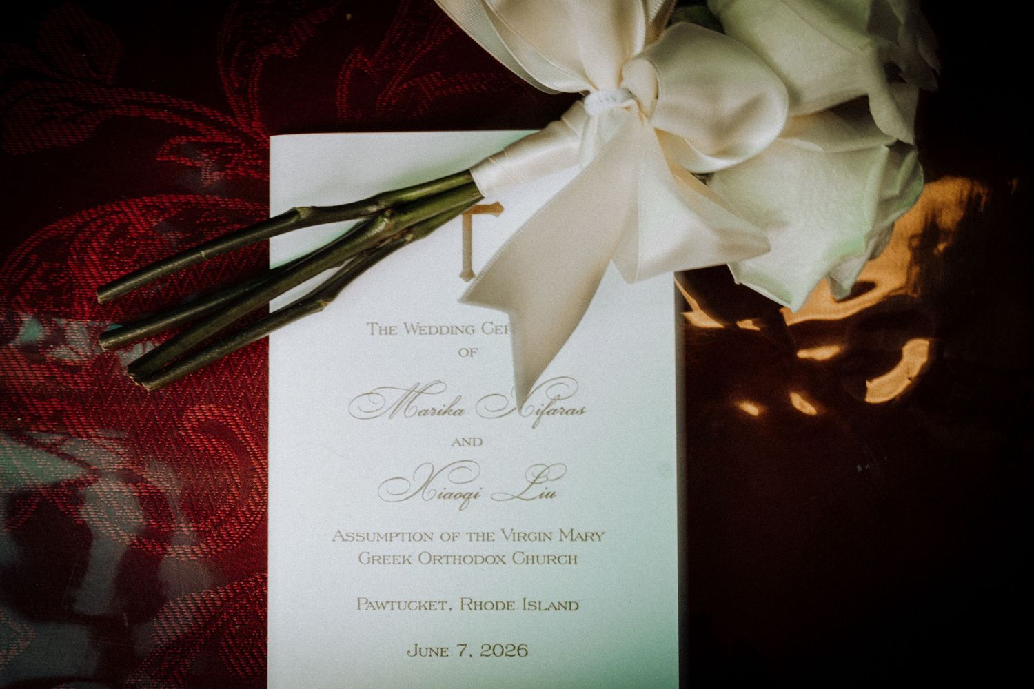 Elegant wedding program with white ribbon bow and white roses on red fabric backdrop.