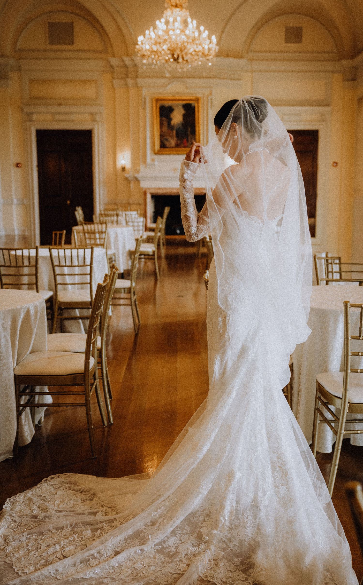 Bride in lace wedding gown with cathedral veil stands in elegant ballroom with gold chiavari chairs and chandelier.