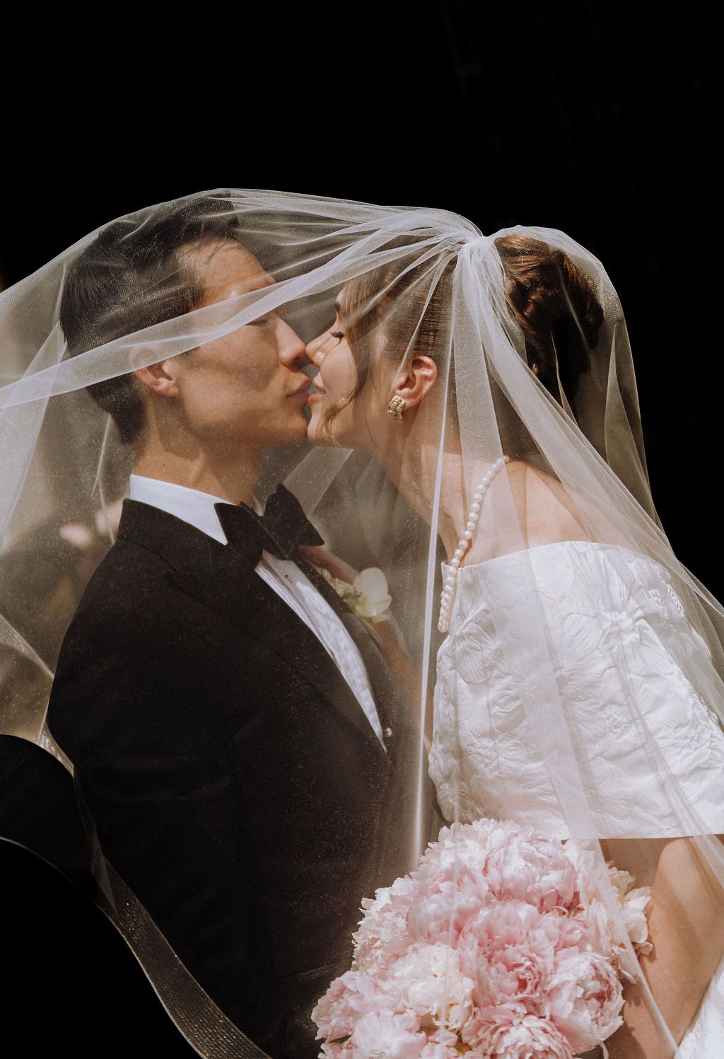 Bride and groom sharing a romantic kiss under a flowing white veil, bride holding pink peonies bouquet.