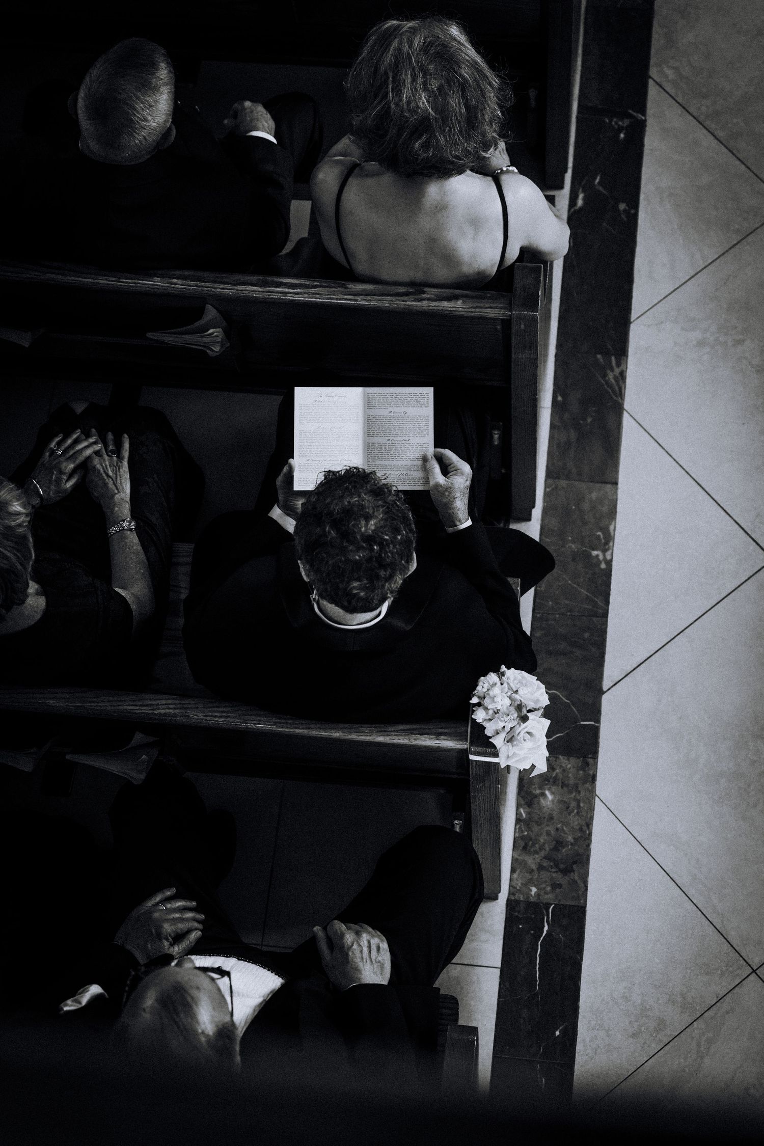 Aerial black and white photo of wedding guests seated in church pews, one holding a bouquet.