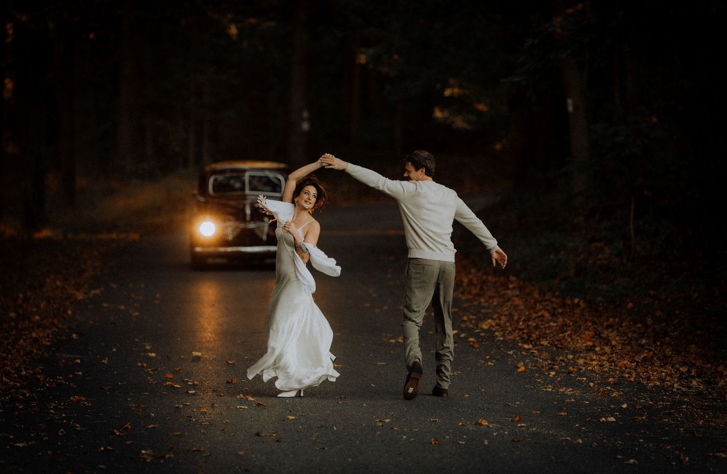 Bride in a white gown and groom in light shirt and trousers dance on a leaf-covered forest road beside a vintage car at dusk.