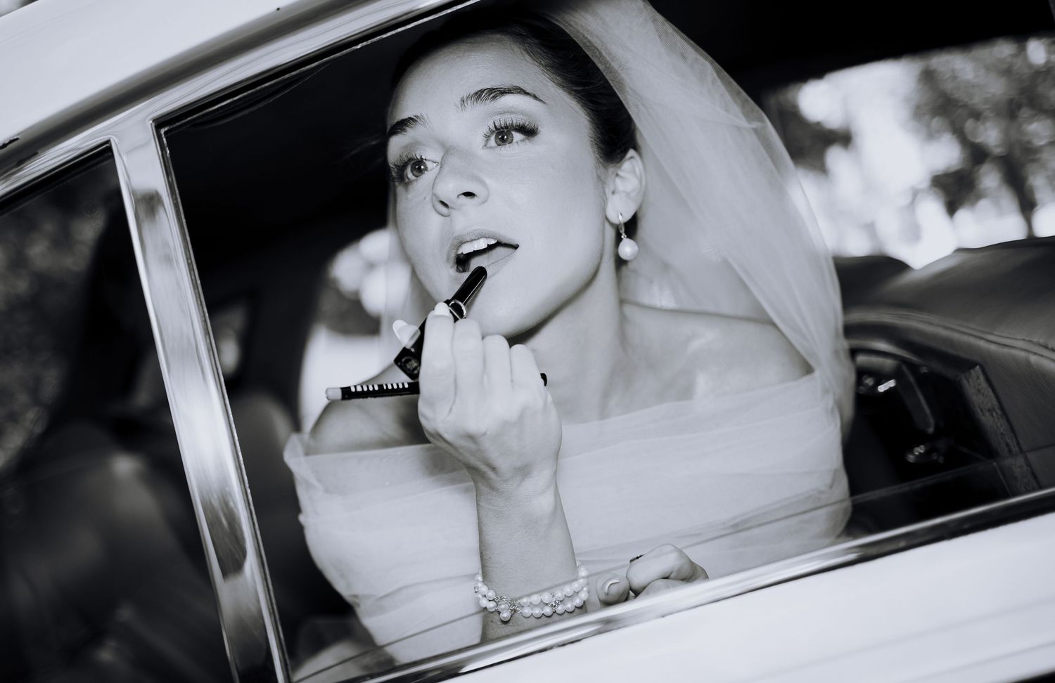 Black and white photo of a bride applying lipstick in a car window on her wedding day.