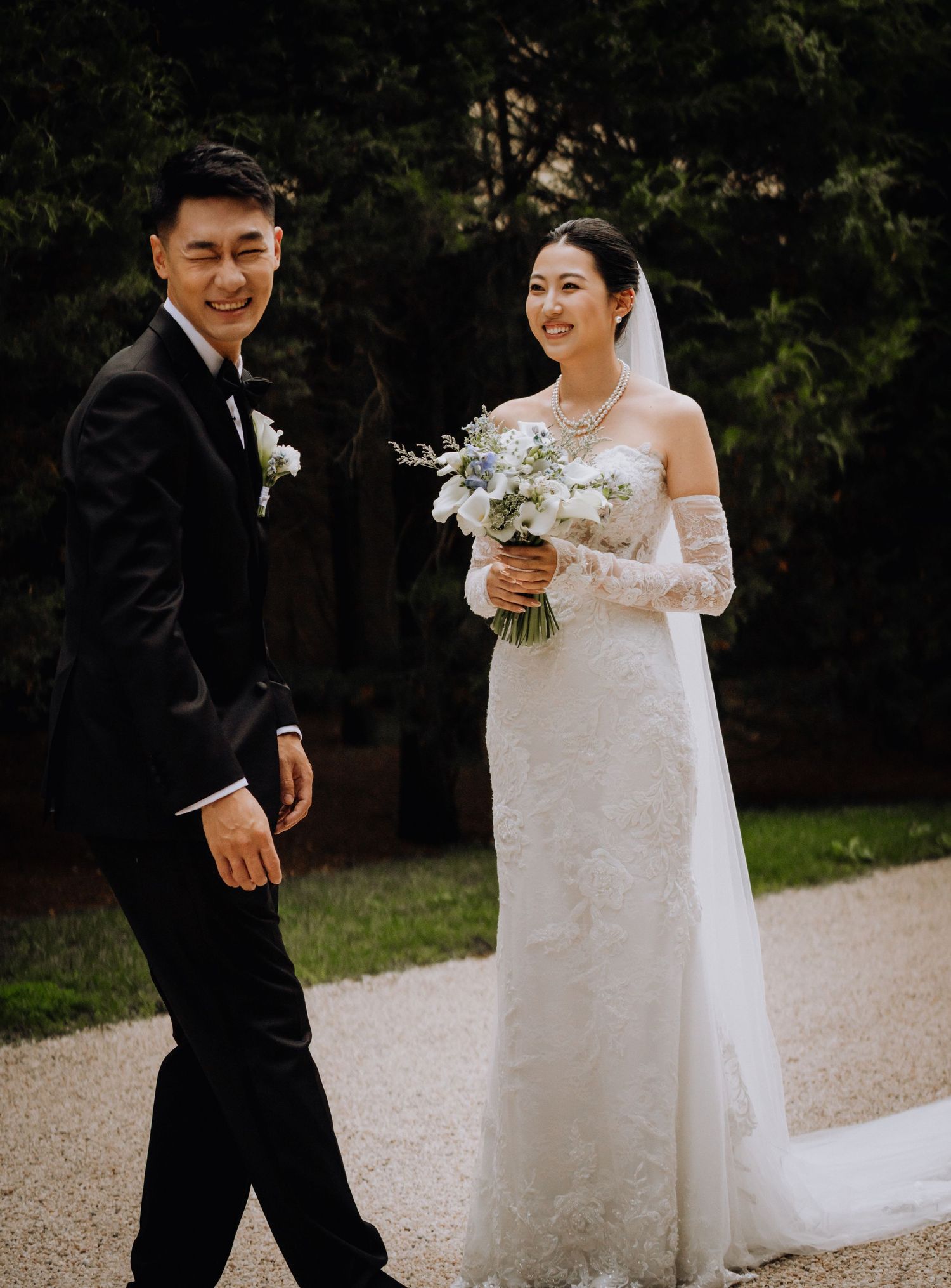 A smiling couple in wedding attire walks outdoors, the bride holding a white bouquet in a lace gown, groom in a black tuxedo.