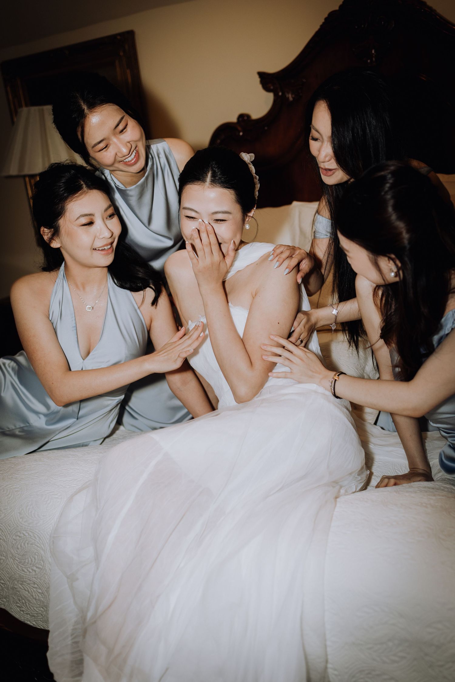 Bride in white gown laughing with bridesmaids in silver dresses on a bed during wedding preparations.