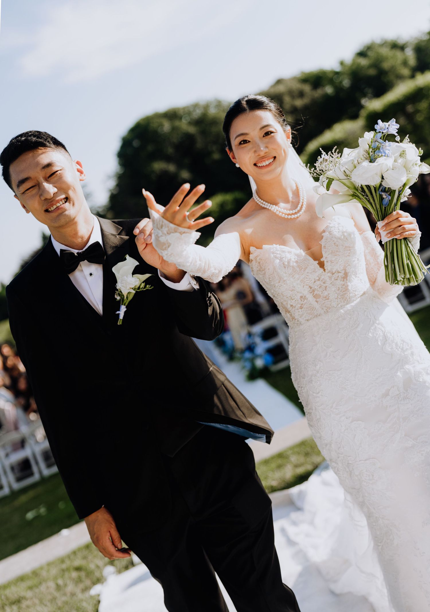 Joyful newlyweds walk down the aisle, bride in lace gown holding white bouquet, groom in tuxedo, showing wedding rings.
