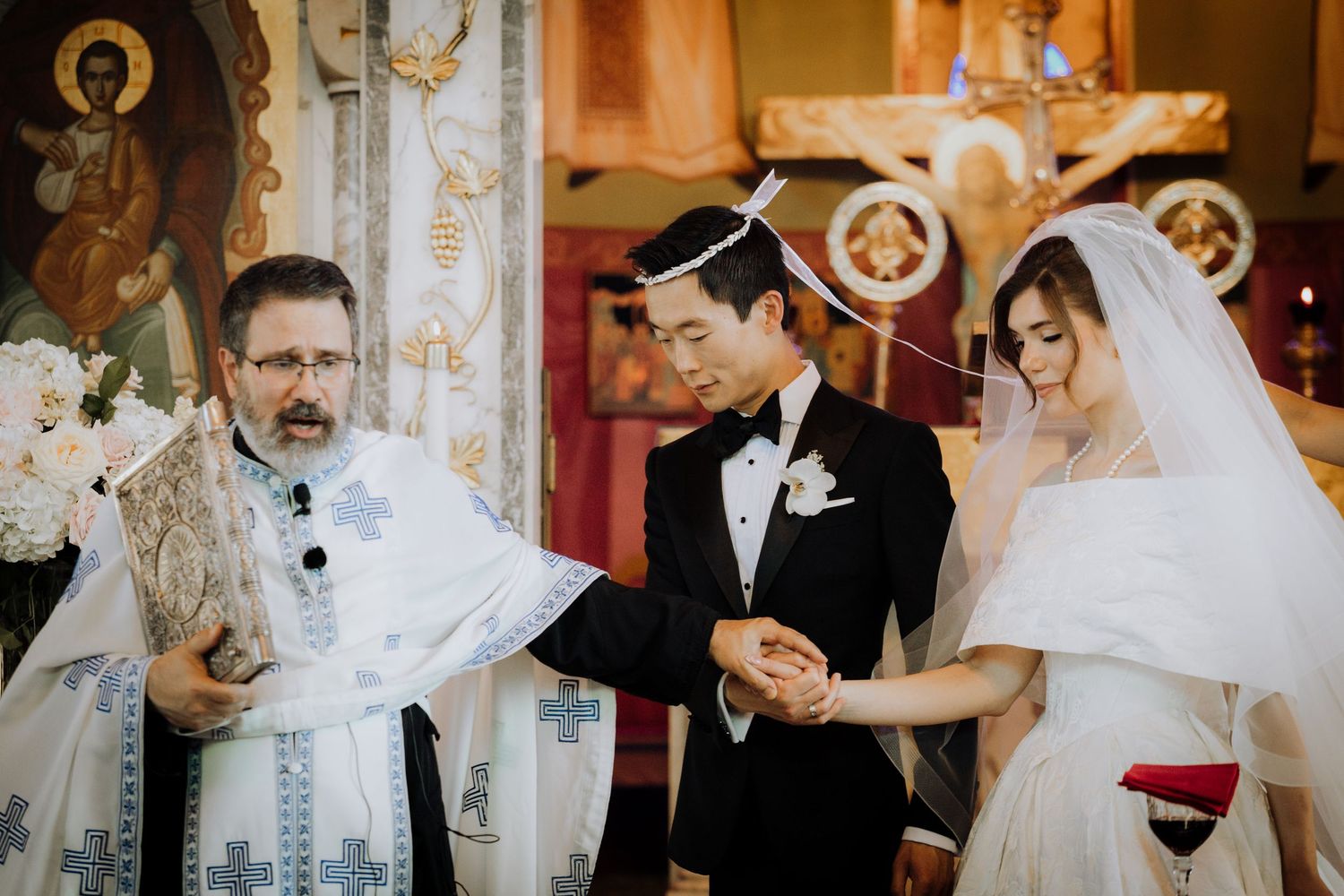 Orthodox priest officiates wedding ceremony joining couple's hands in ornate church with bride in white veil and groom in tuxedo.