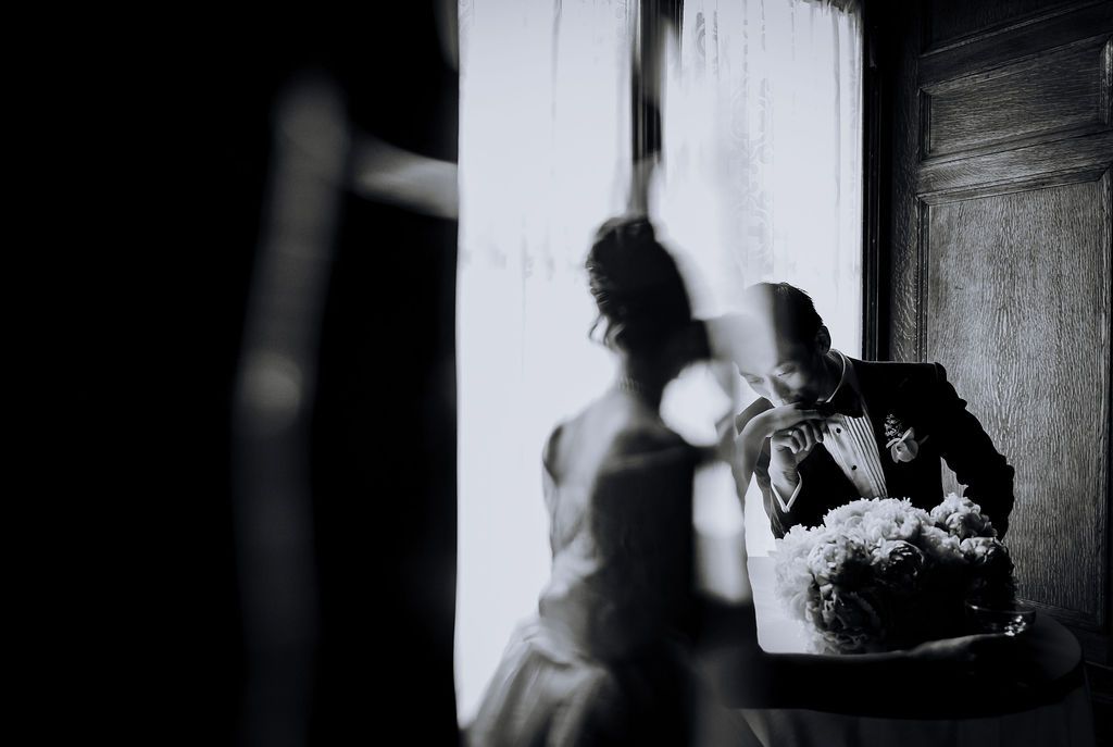 Black and white wedding photo of bride and groom near window, groom smelling bouquet of white flowers.