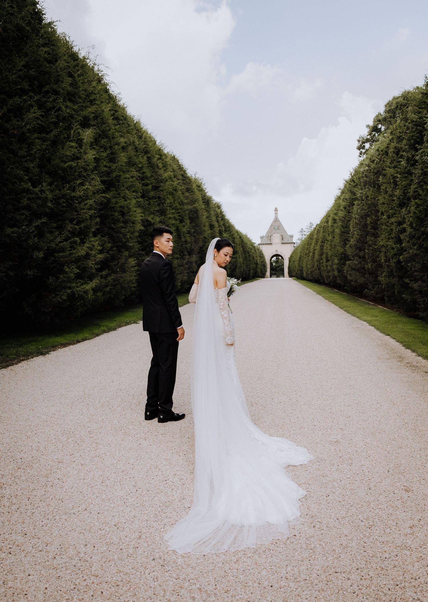 Bride and groom stand on a grand gravel path lined with tall hedges, her long white veil trailing elegantly behind.