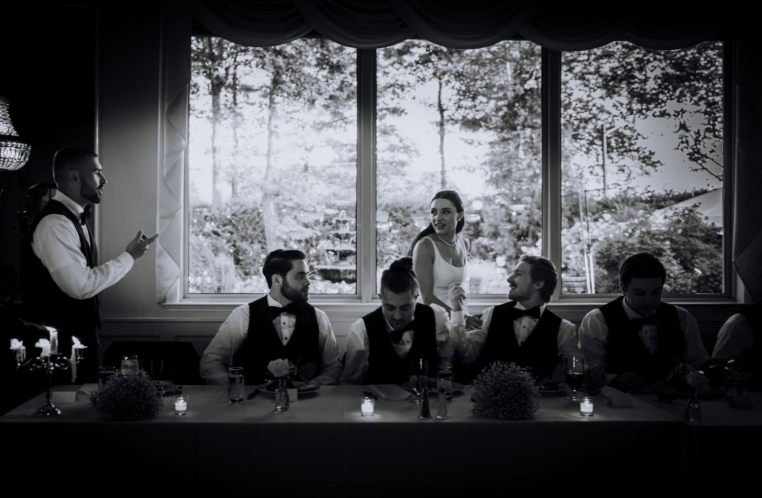 Black and white photo of wedding party seated at a long table with scenic window views.