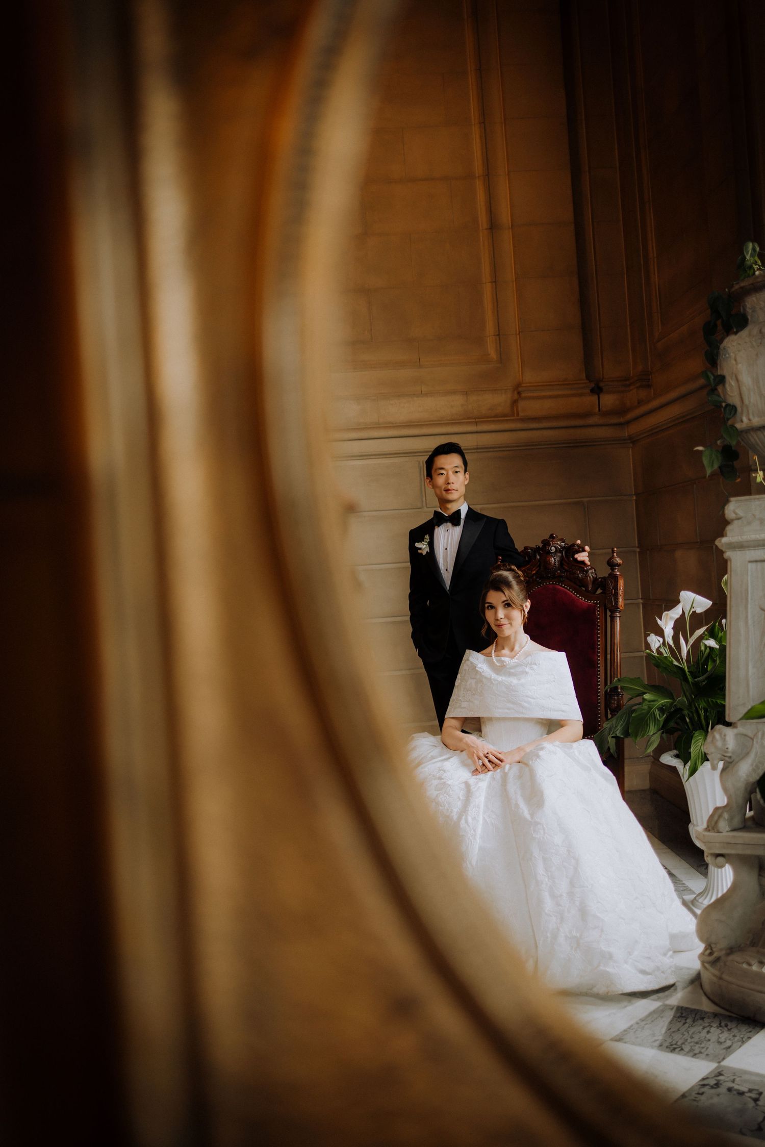 Bride in white gown and groom in black tuxedo pose elegantly in ornate hall, reflected in vintage gold mirror.