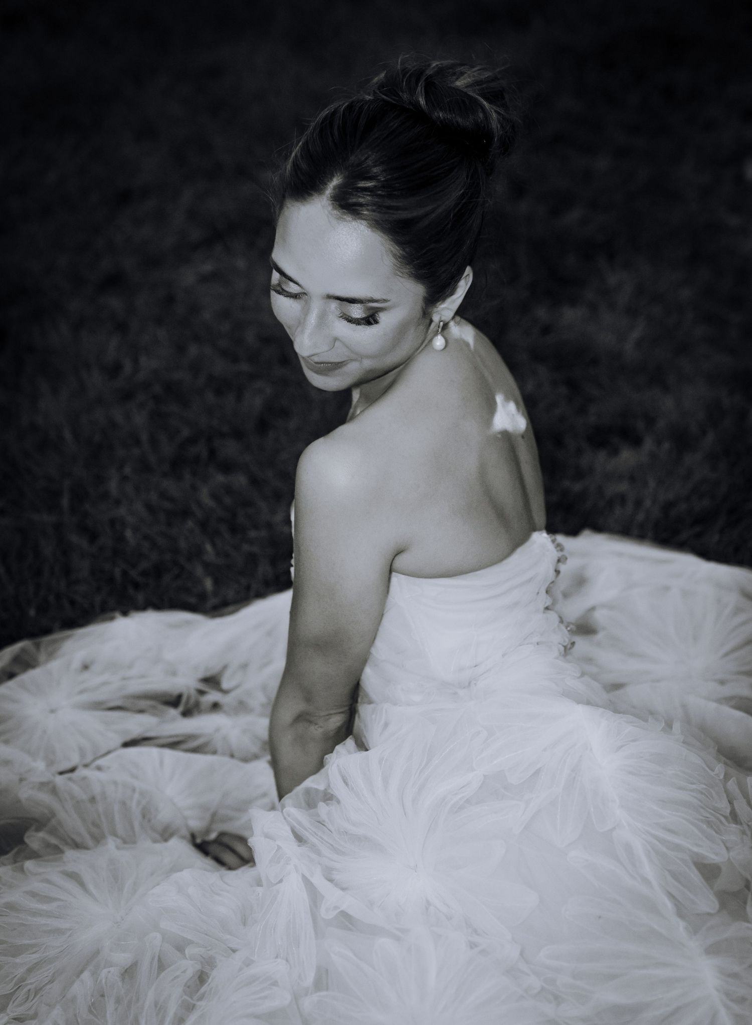 Black and white photo of a bride in a strapless tulle gown sitting gracefully on grass, looking down elegantly.