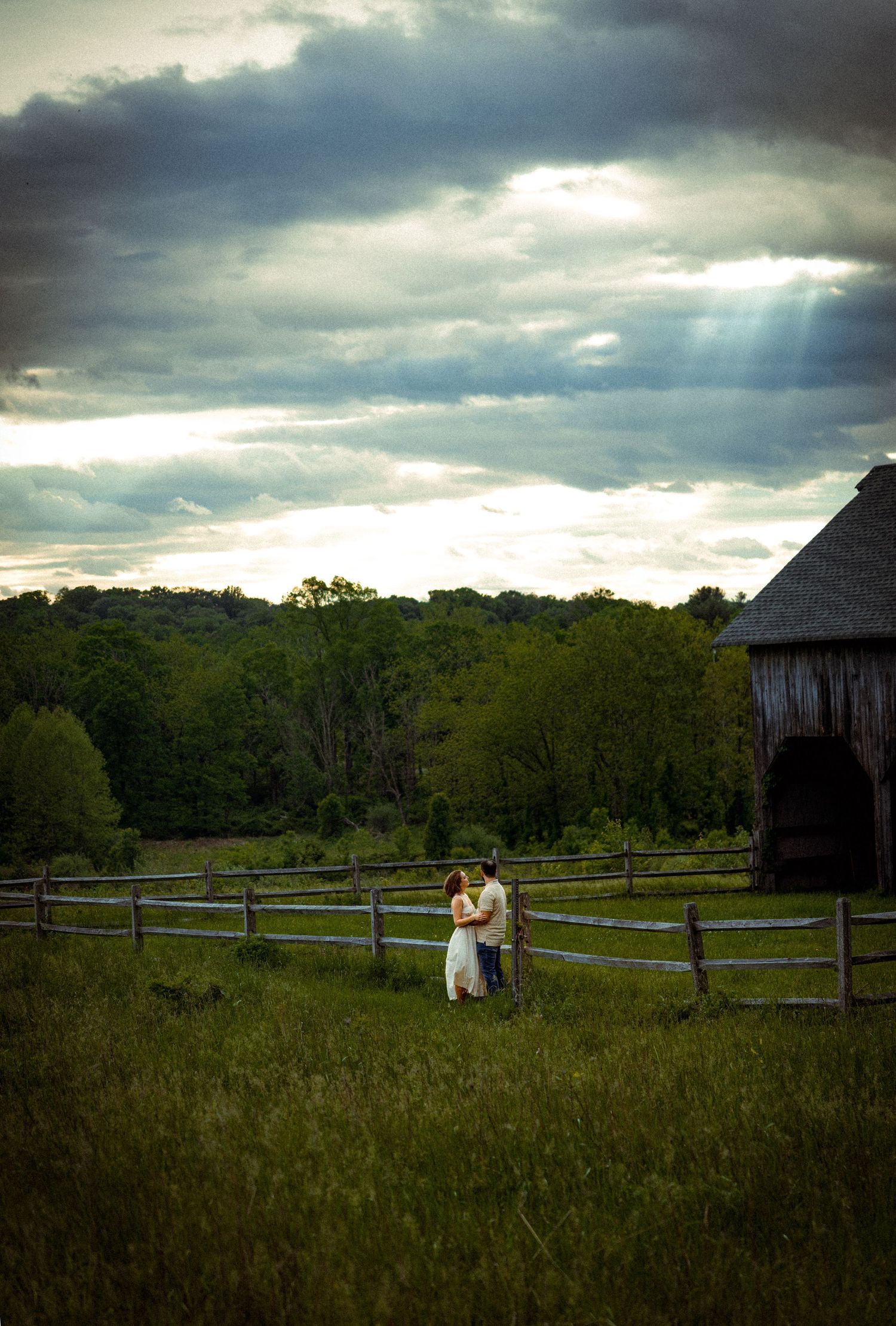 A couple sits in a rustic field with dramatic cloudy skies and wooden fencing.