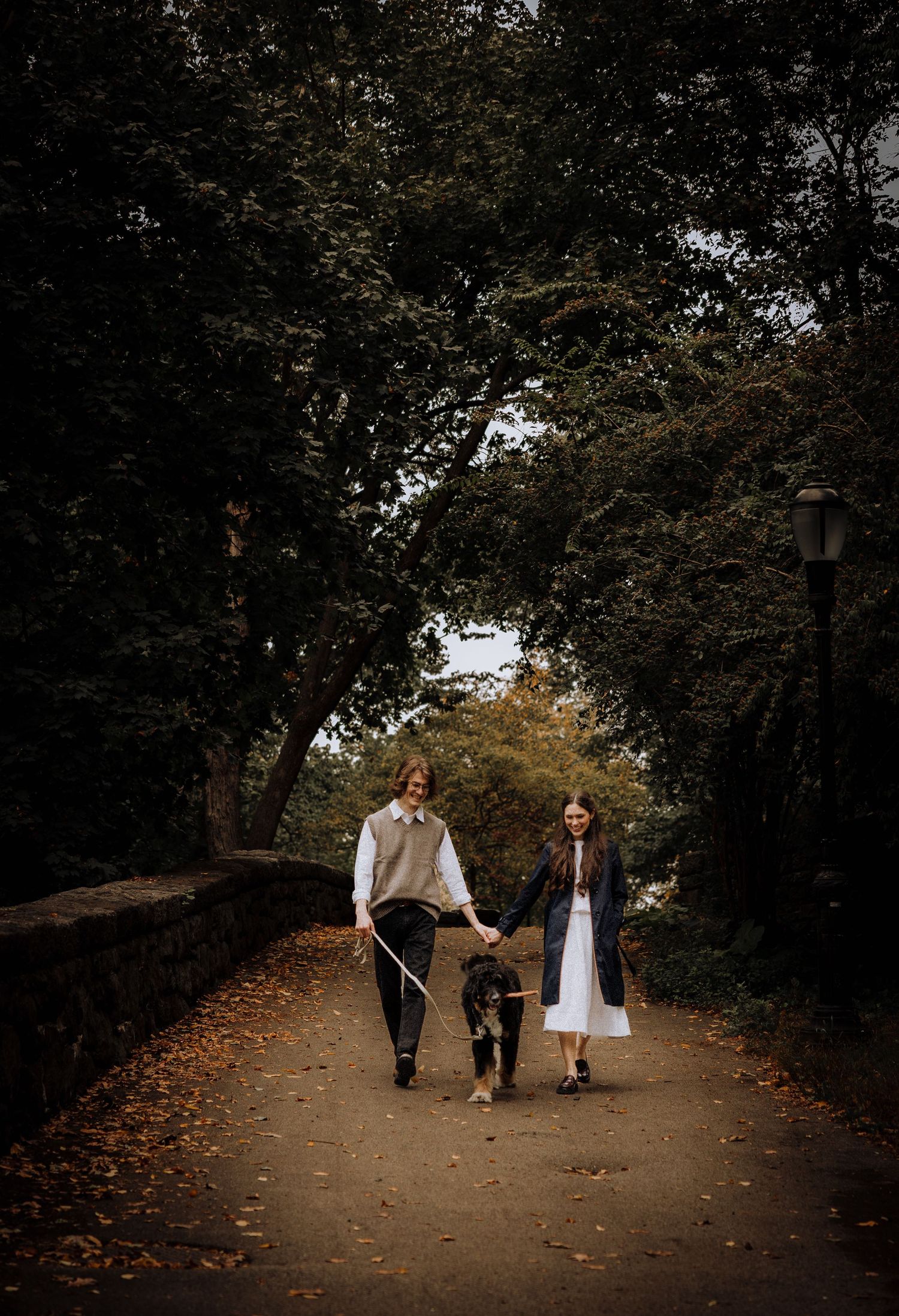 People stroll down a tree-lined path covered in fallen autumn leaves.