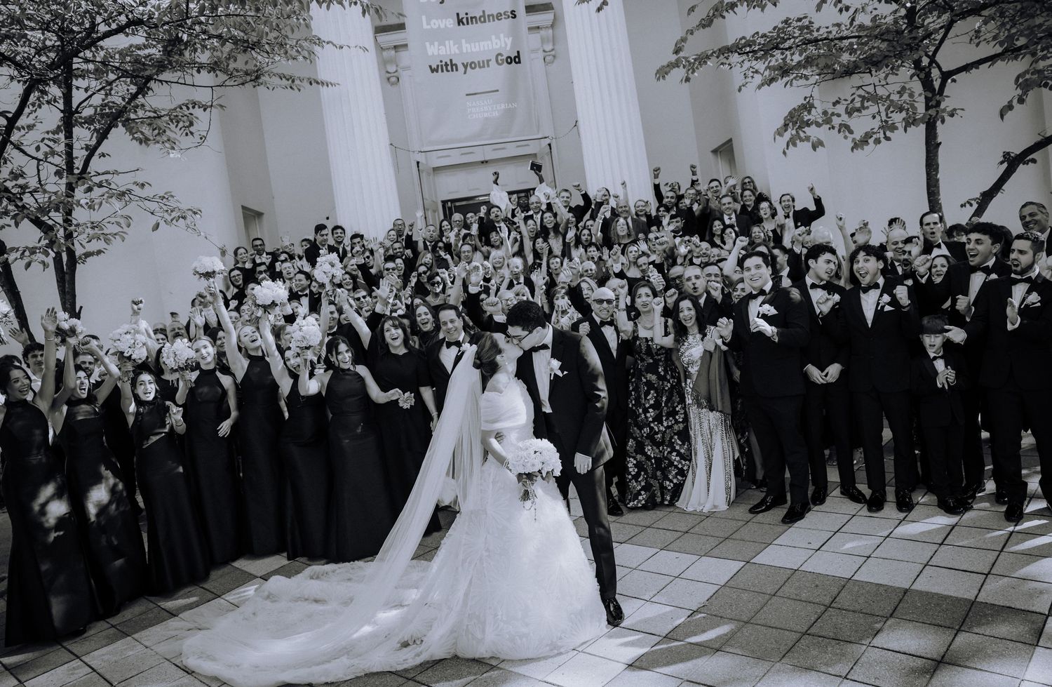 Black and white wedding photo of bride and groom with large wedding party on grand steps, guests cheering.